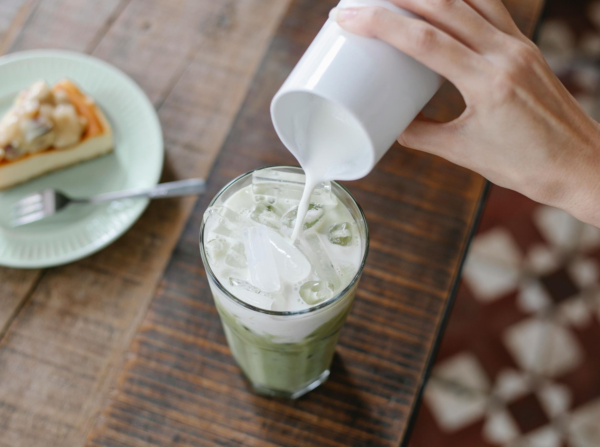 Hand pouring milk into an iced matcha latte, wooden table, slice of cake on a plate.
