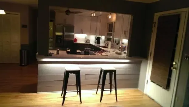 Two bar stools in front of a pass-through to a lit kitchen with white cabinets, dark wall.