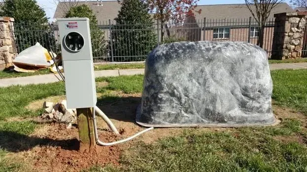 Outdoor electrical box next to a large, rock-shaped utility cover on grass. A fence and houses are in the background.