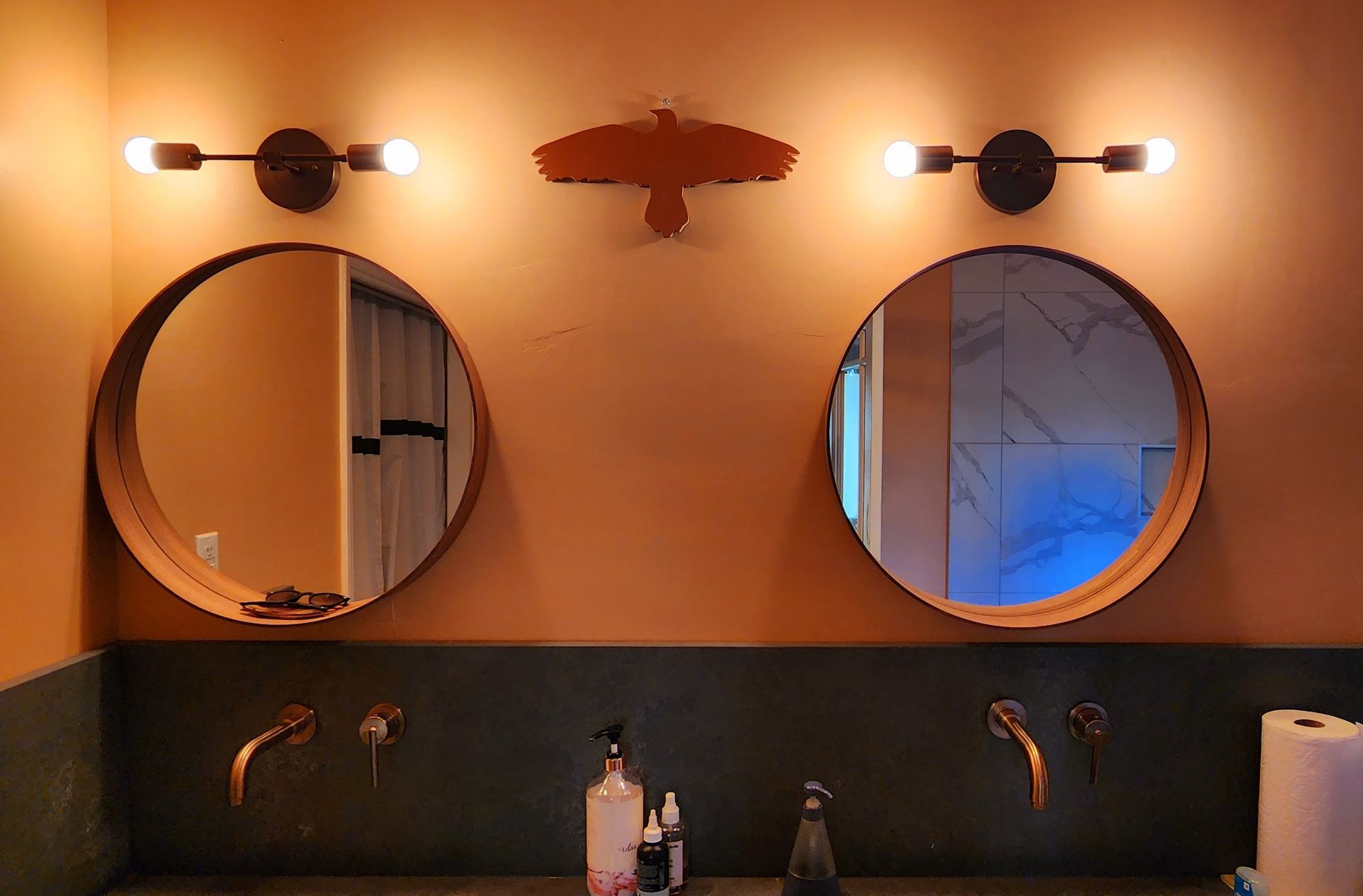 Bathroom with two round mirrors, copper faucets, and sconce lighting on a salmon-colored wall.