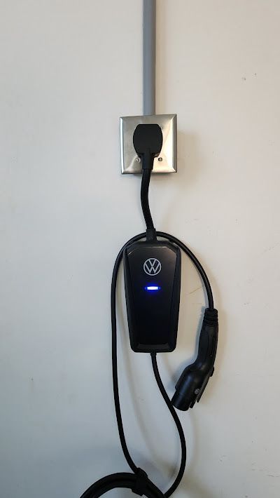 Black electric vehicle charger plugged into a wall outlet, mounted on a white wall.