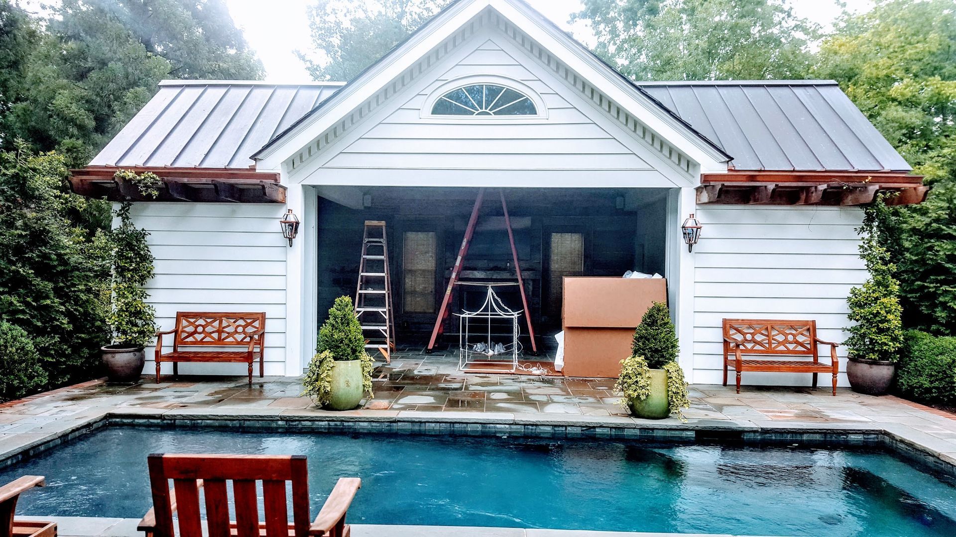 Poolside cottage with open garage, ladders, and furniture. White exterior, blue pool.