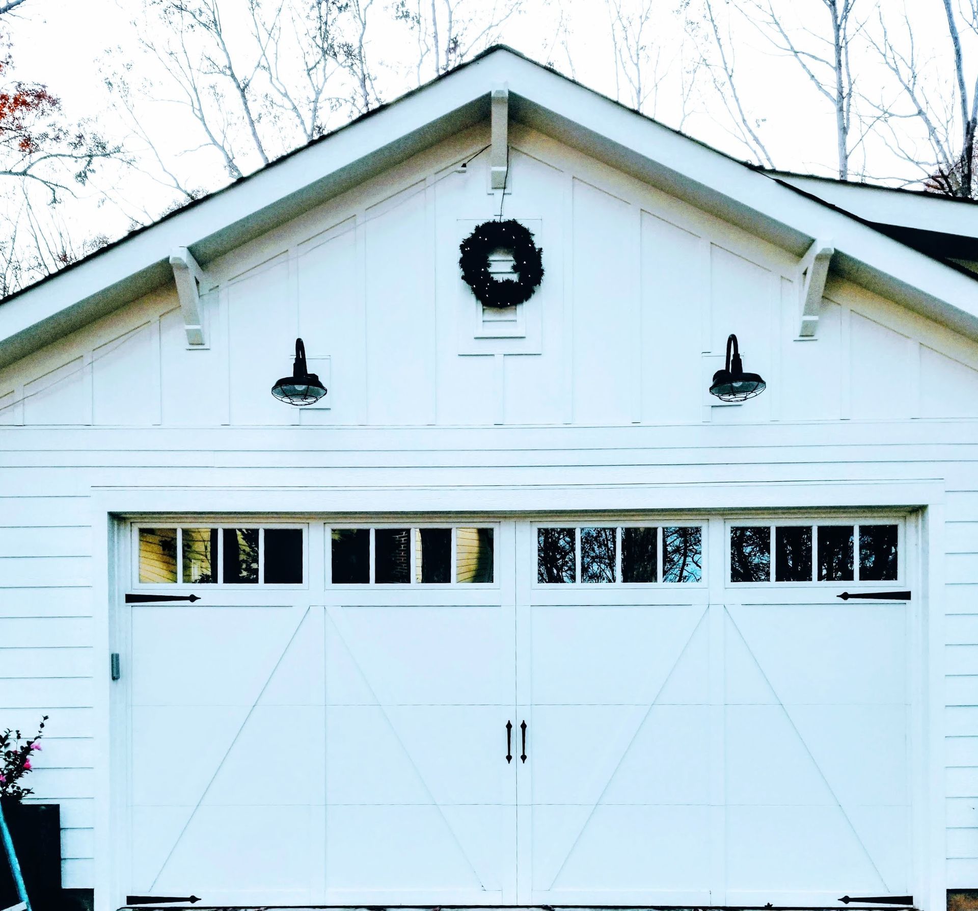White garage with two doors, glass windows, black hardware, and sconce lights. A wreath hangs at the top.