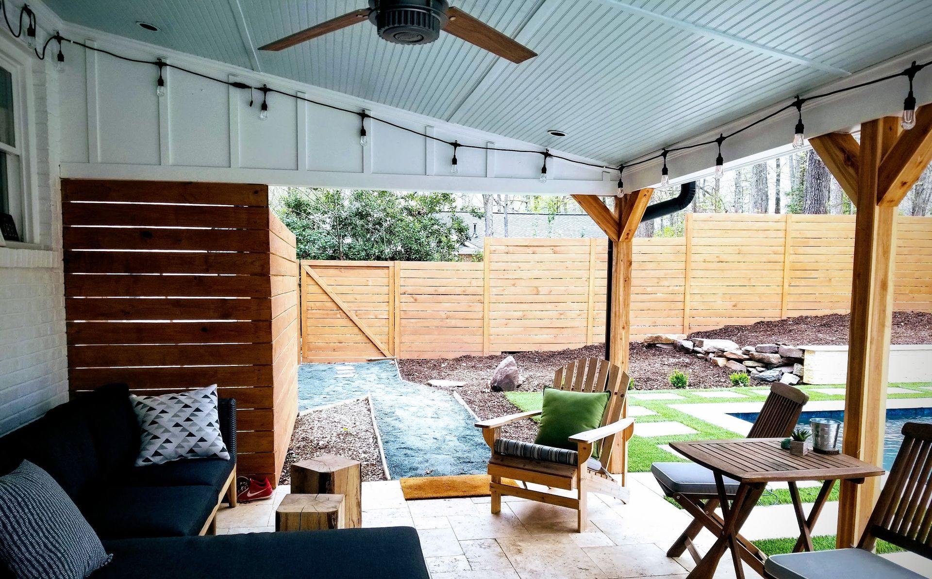 Outdoor patio with seating, wooden accents, string lights, and a view of a backyard with a pool.