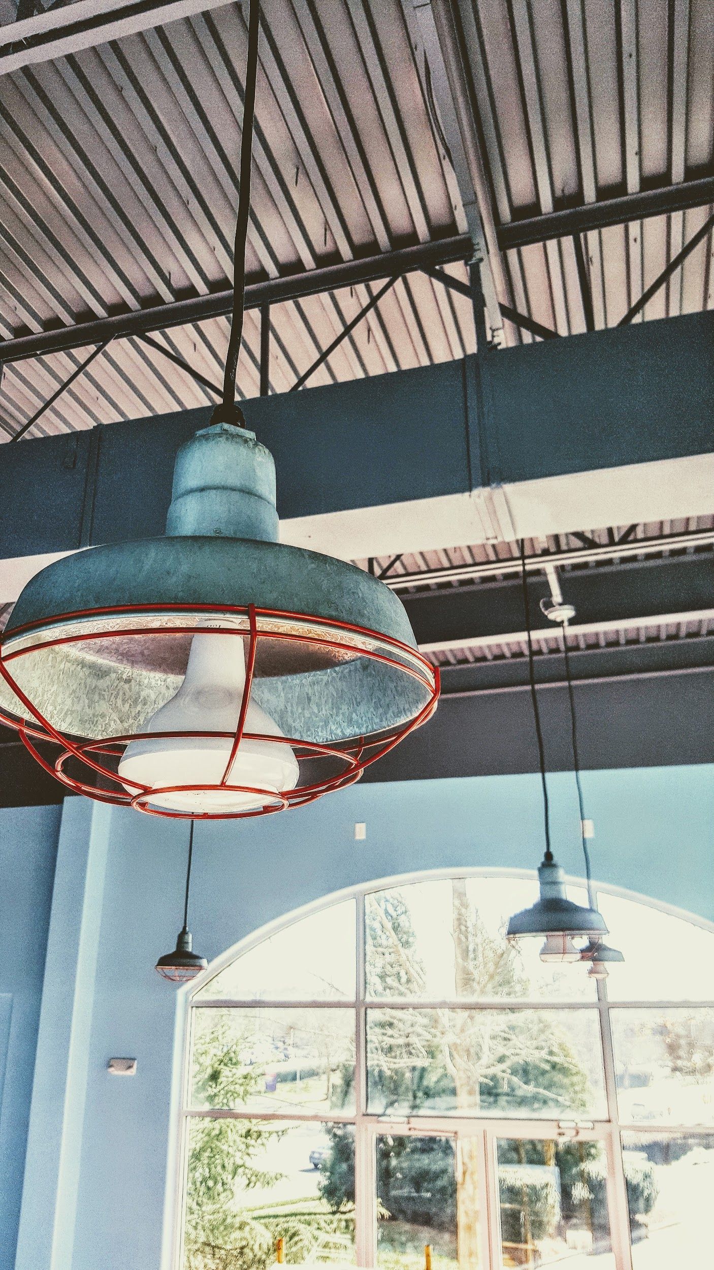 Industrial-style hanging light fixtures with metal cages, against a high, blue-painted ceiling and windows.