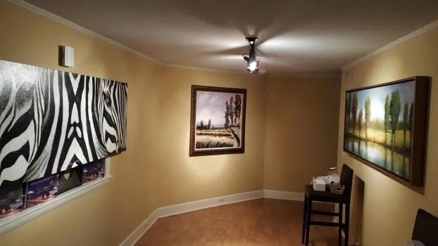 Gallery space with three framed artworks, zebra print, landscape, and shoreline. Beige walls, wood floor, track lighting.