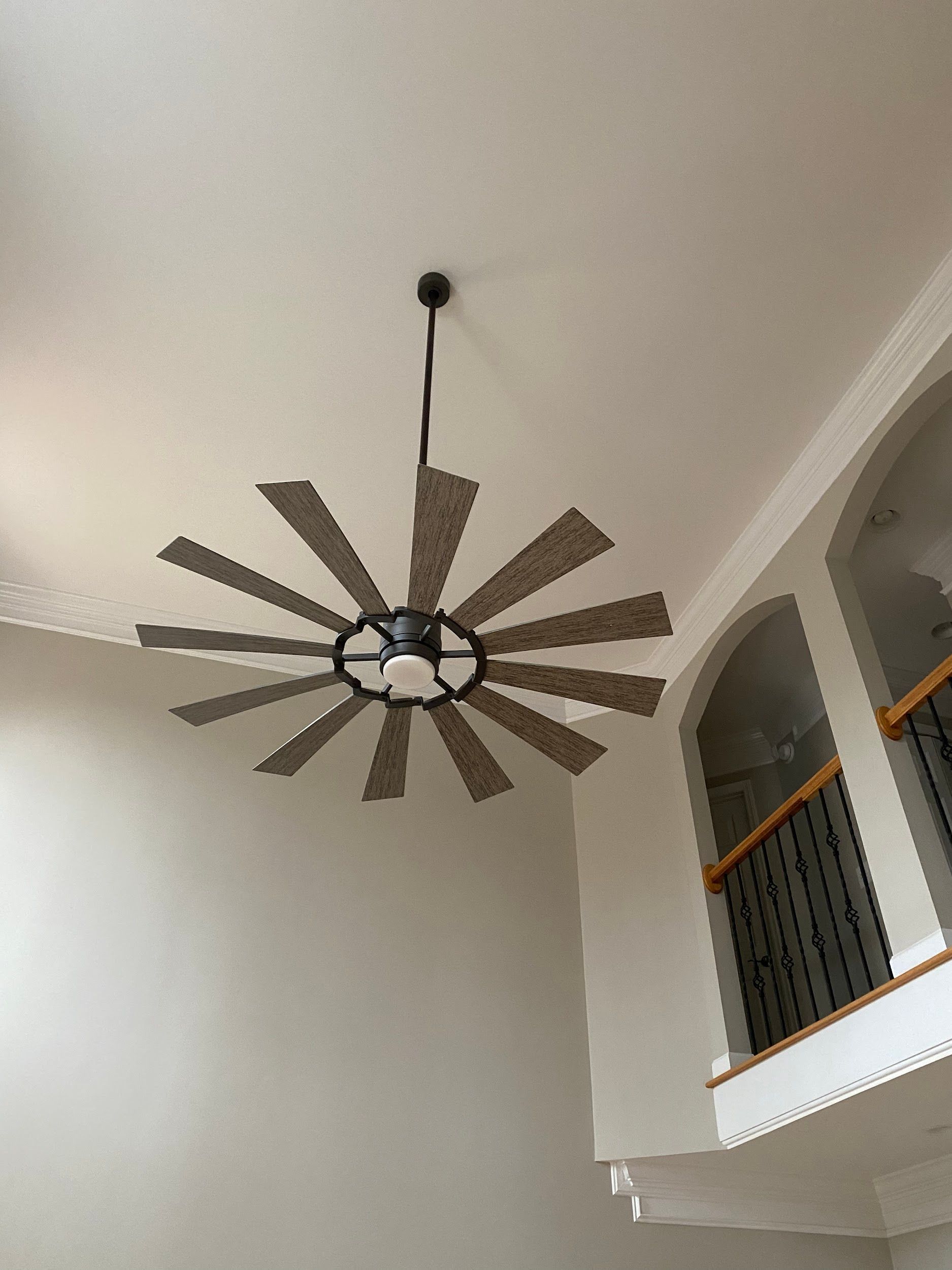 Ceiling fan with wooden blades in a room with a high ceiling and a second-floor balcony.