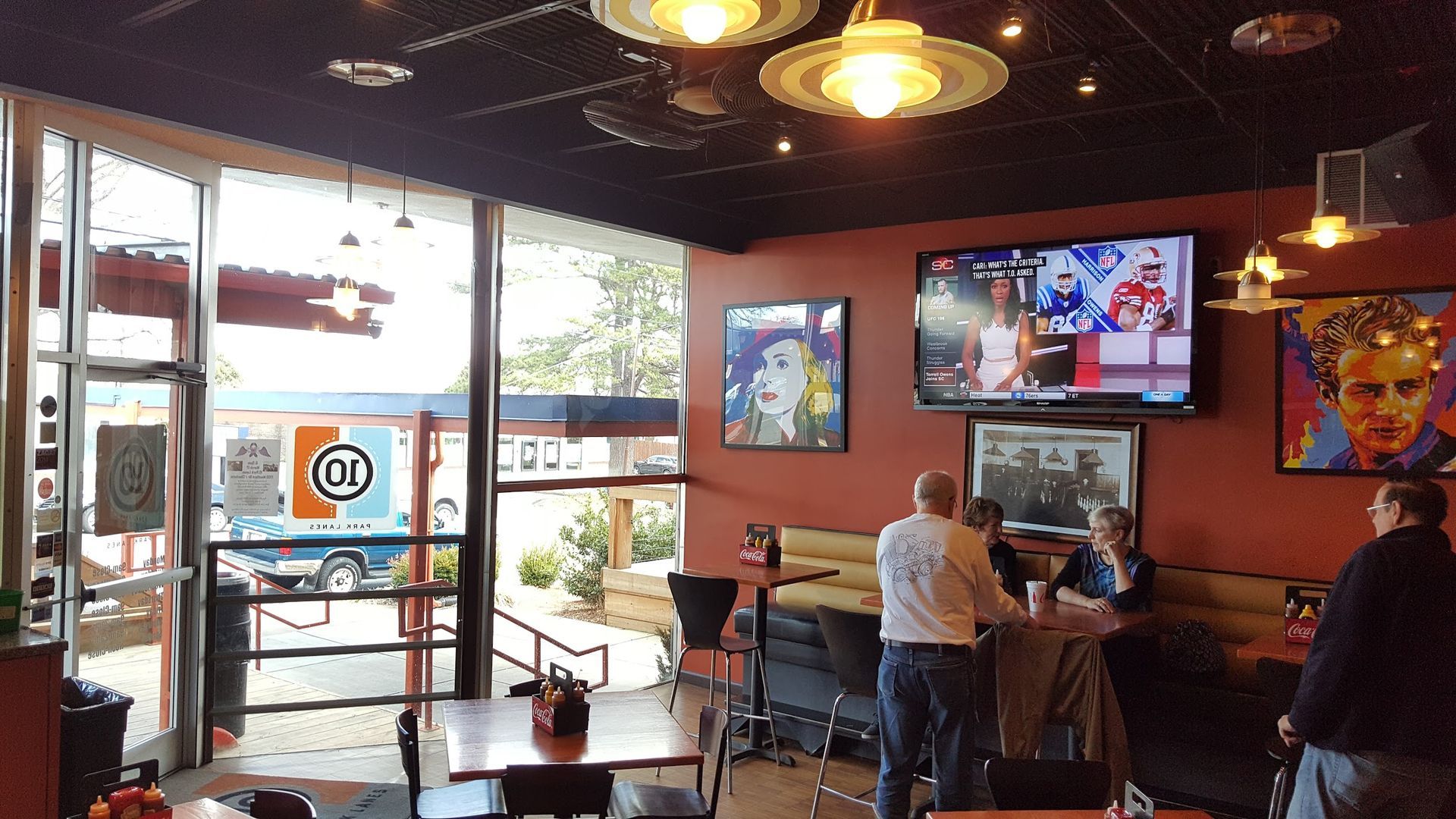 Interior of a restaurant with a TV showing sports, patrons seated at tables, and a view of the outside through a glass door.
