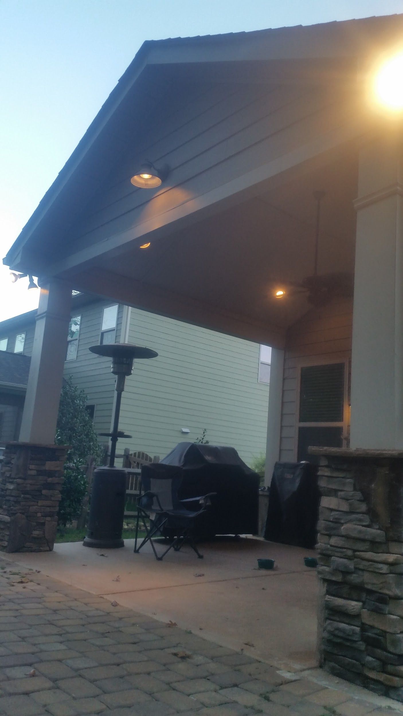 Covered patio with a grill, heater, and built-in lights. Brown roof, stone pillars, and a green building in the background.
