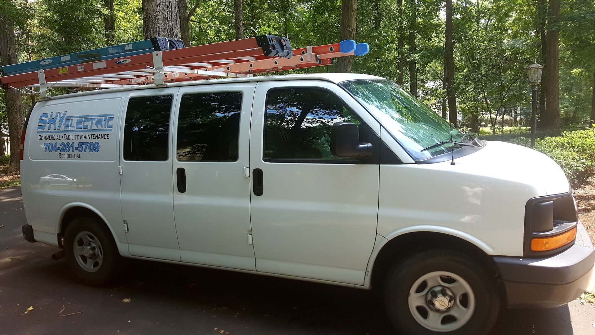 White work van with ladder on roof, parked on a road, trees in background.