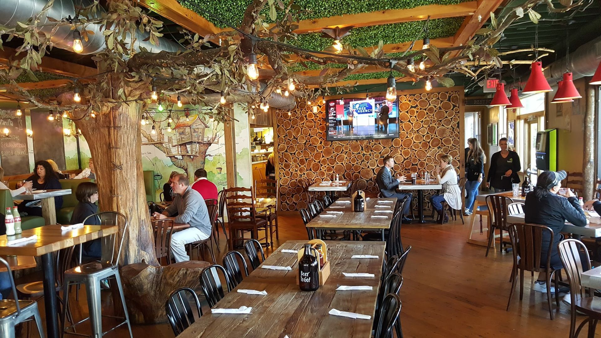 Restaurant interior with rustic decor, wooden tables, tree-like structures, and people dining.