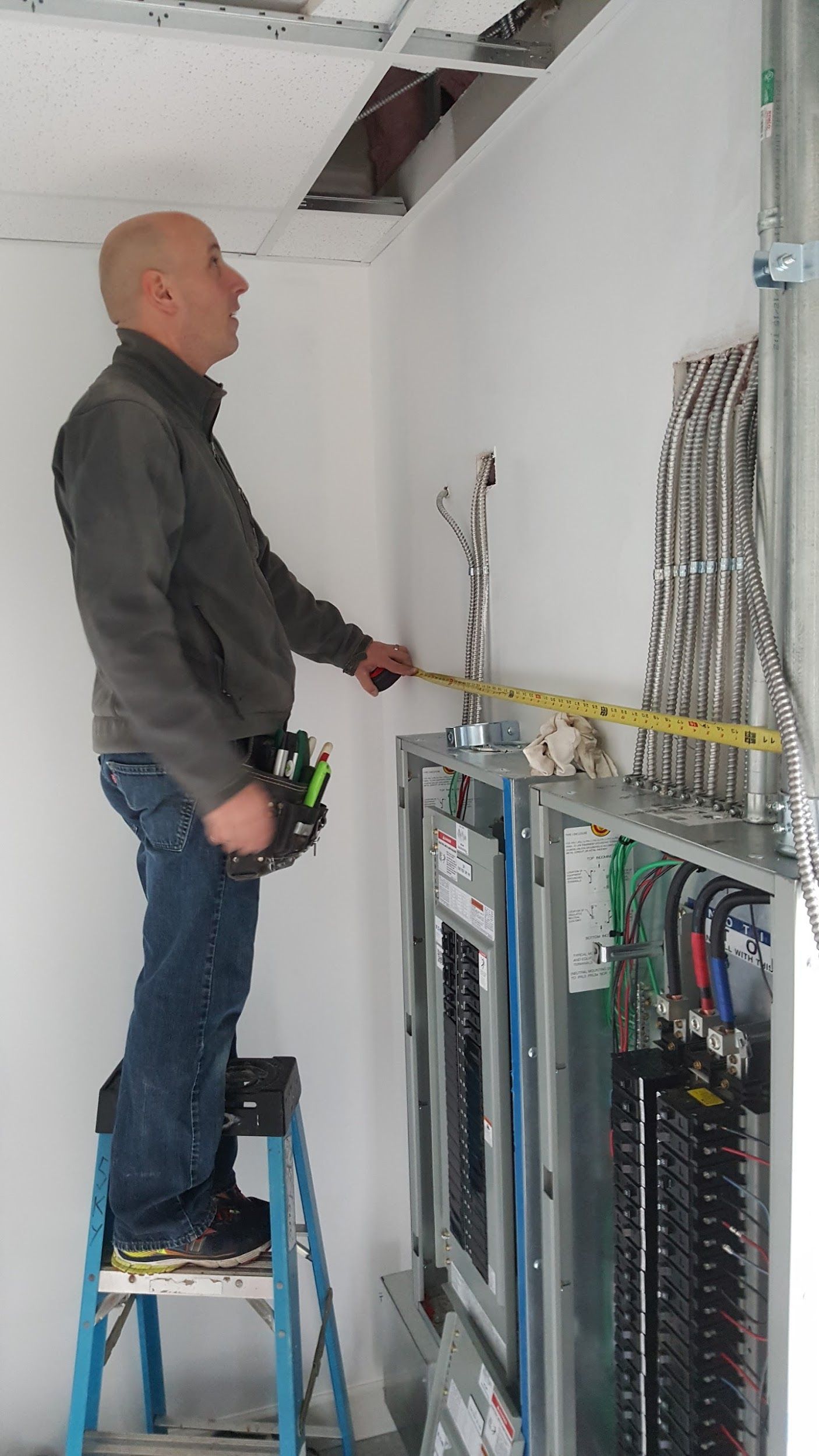 Electrician measuring panel on a ladder in a room with a white wall and an open ceiling.