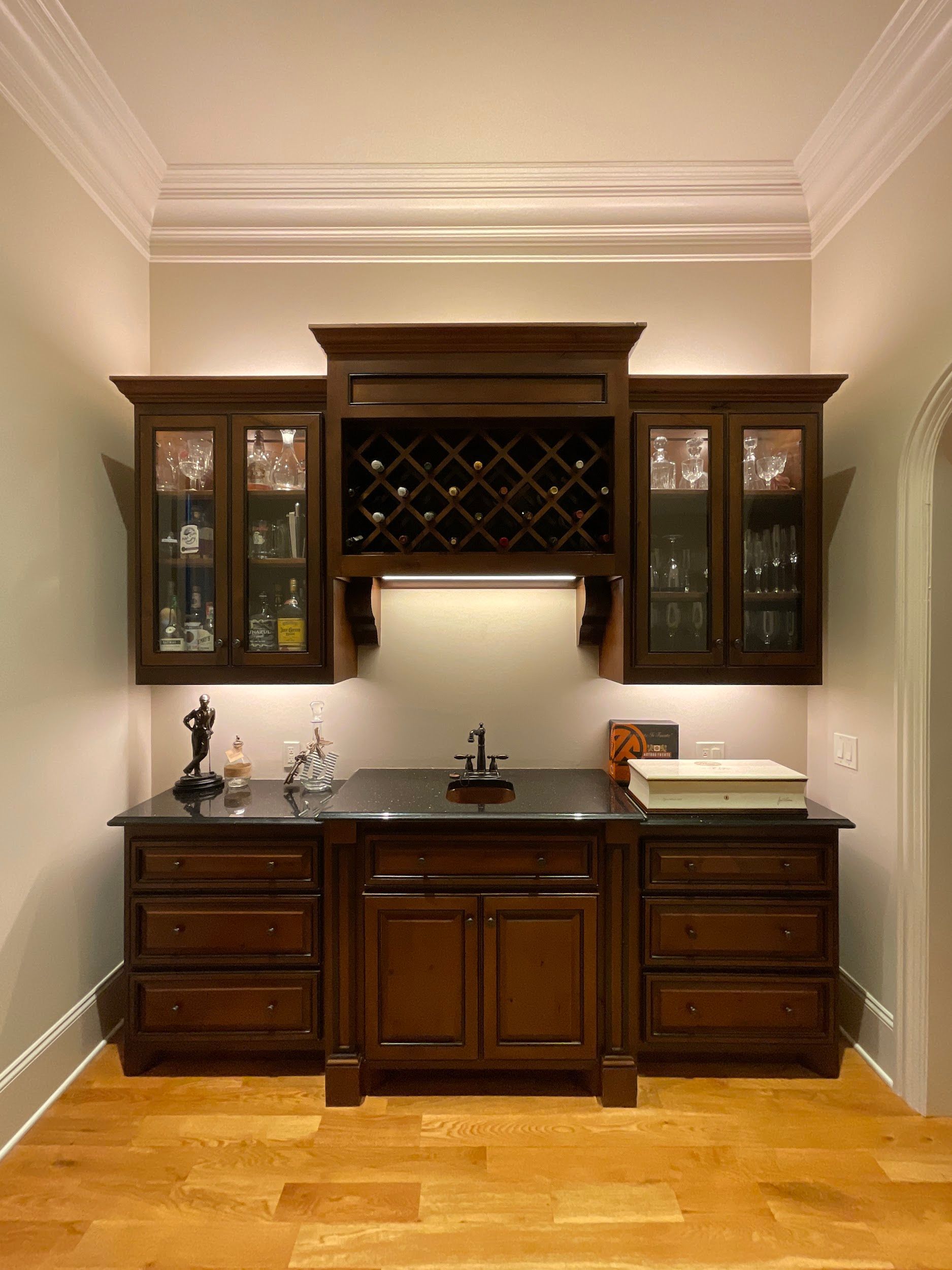 Dark wood home bar with cabinets, a wine rack, and a sink, lit by under-cabinet lights.