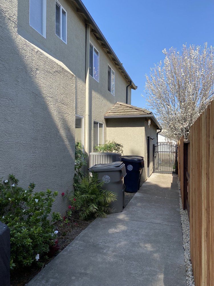 Narrow walkway between two-story building and wooden fence. Trash cans sit near a small structure. Sunny day.