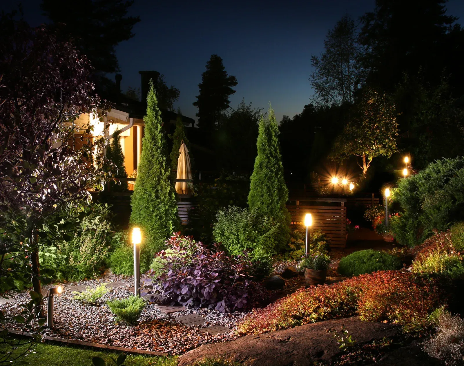 Nighttime garden scene with illuminated plants and pathways near a house.