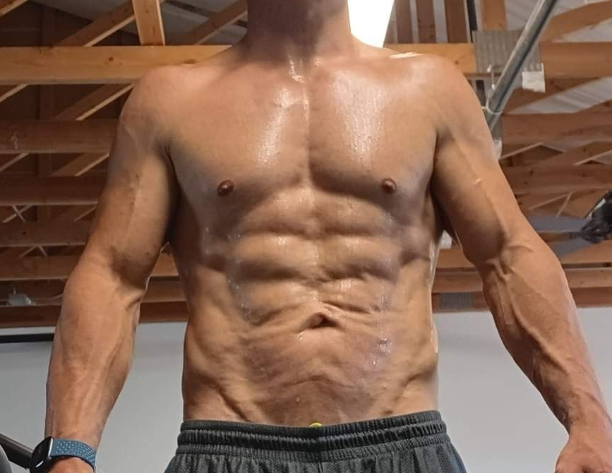 Man with defined abs and arms, shirtless, sweating, in a gym setting.