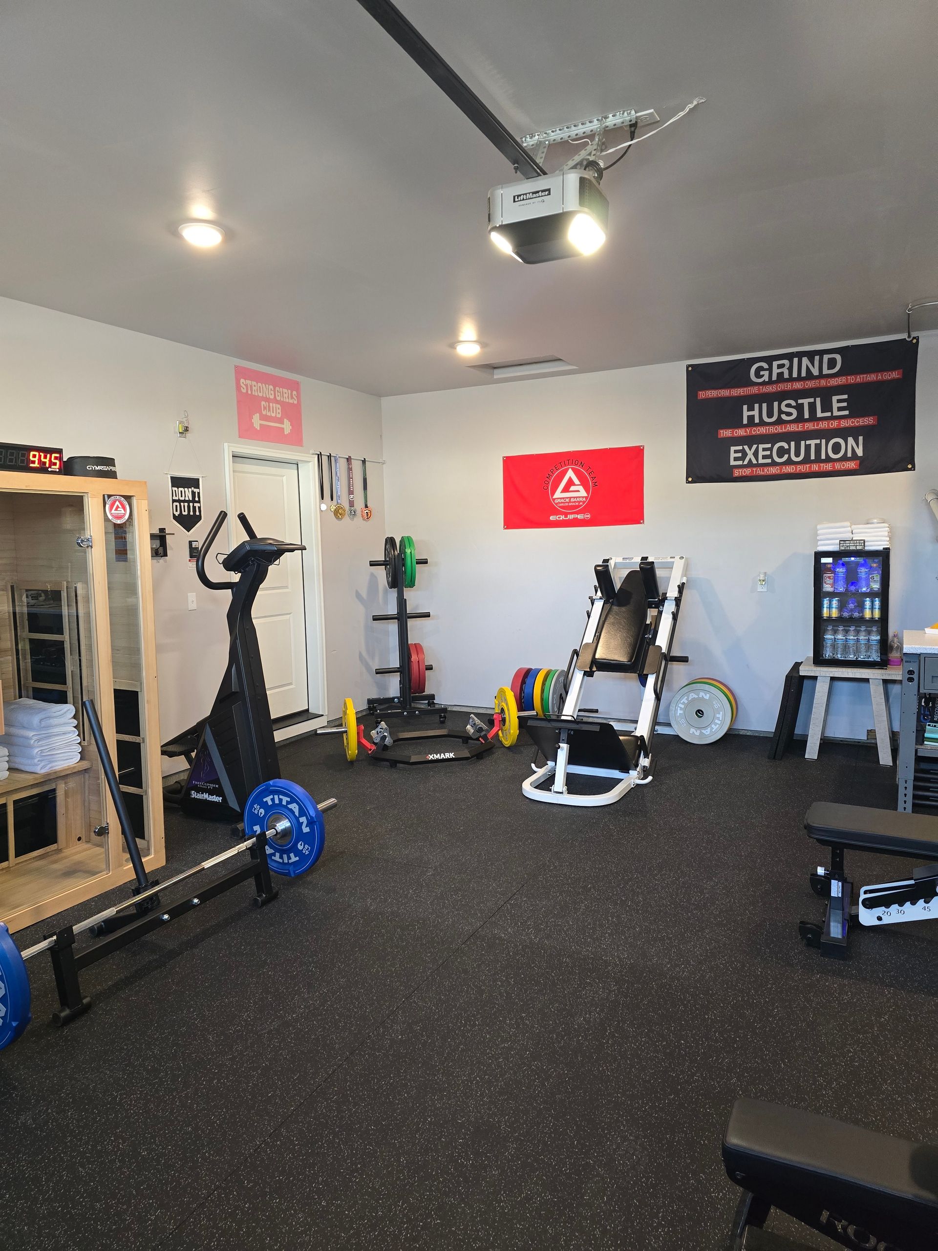 Woman exercising on an air bike in a garage gym. She's reaching up, wearing black athletic clothing.