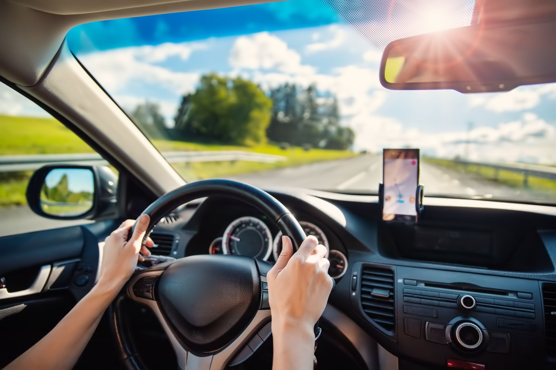 Driver holding steering wheel on a sunny highway, with a phone mounted on the dashboard.