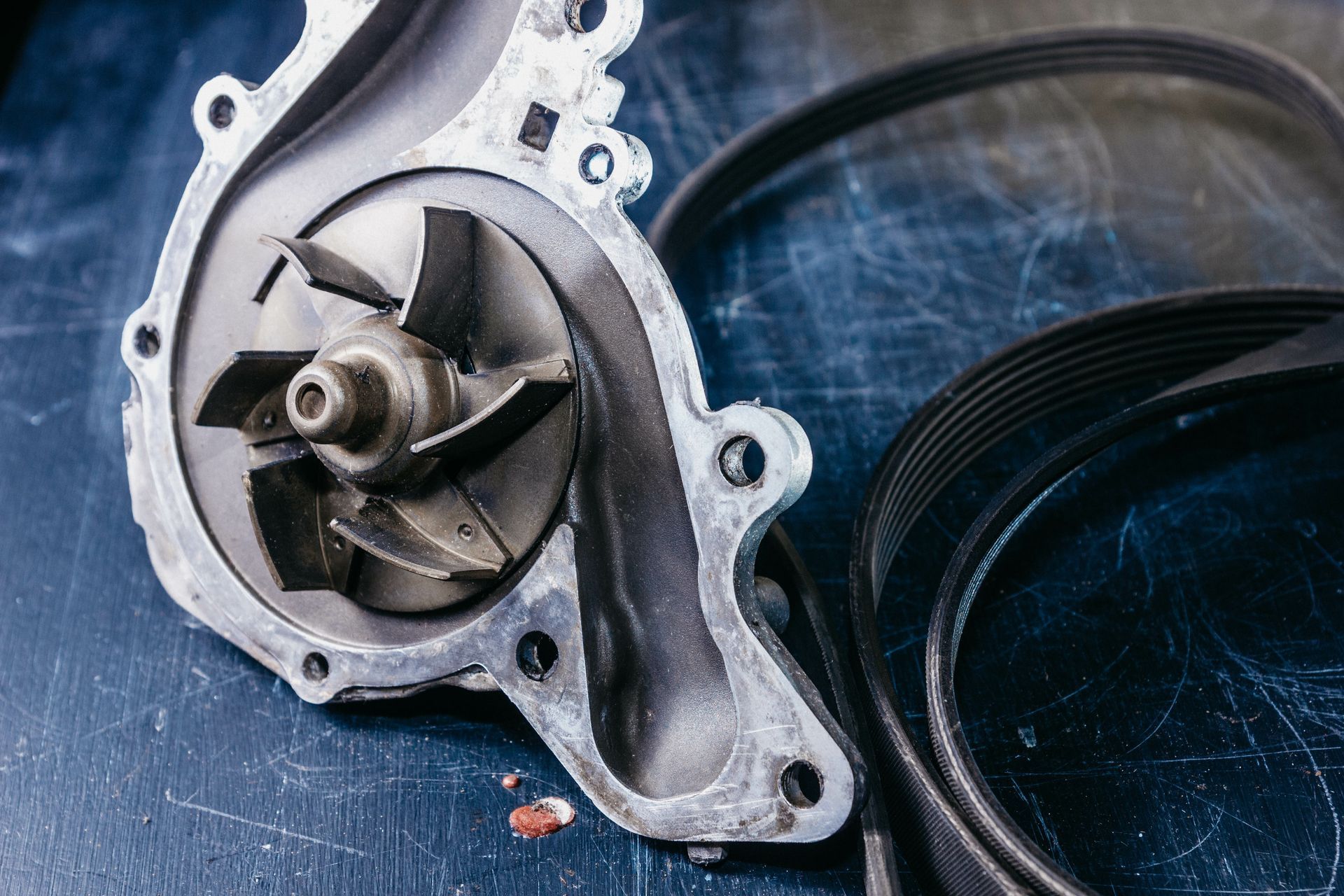 Close-up of a metal water pump housing and impeller beside a black serpentine belt on a blue surface