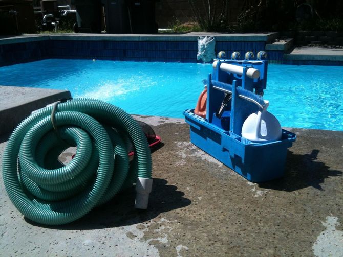 Pool cleaning supplies beside a blue swimming pool: coiled green hose and blue caddy on the deck.