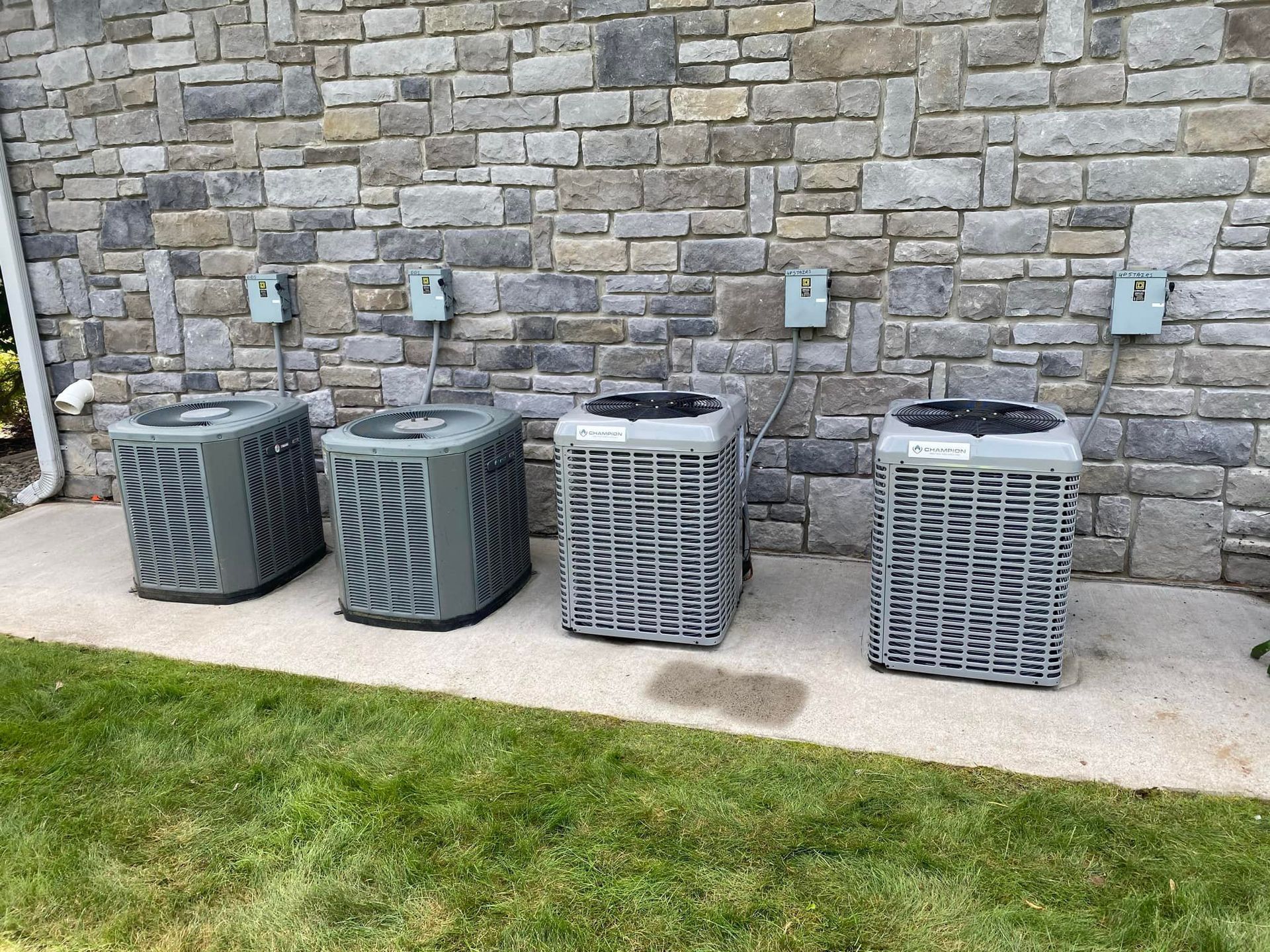 Three air conditioners are sitting on a sidewalk in front of a brick wall.