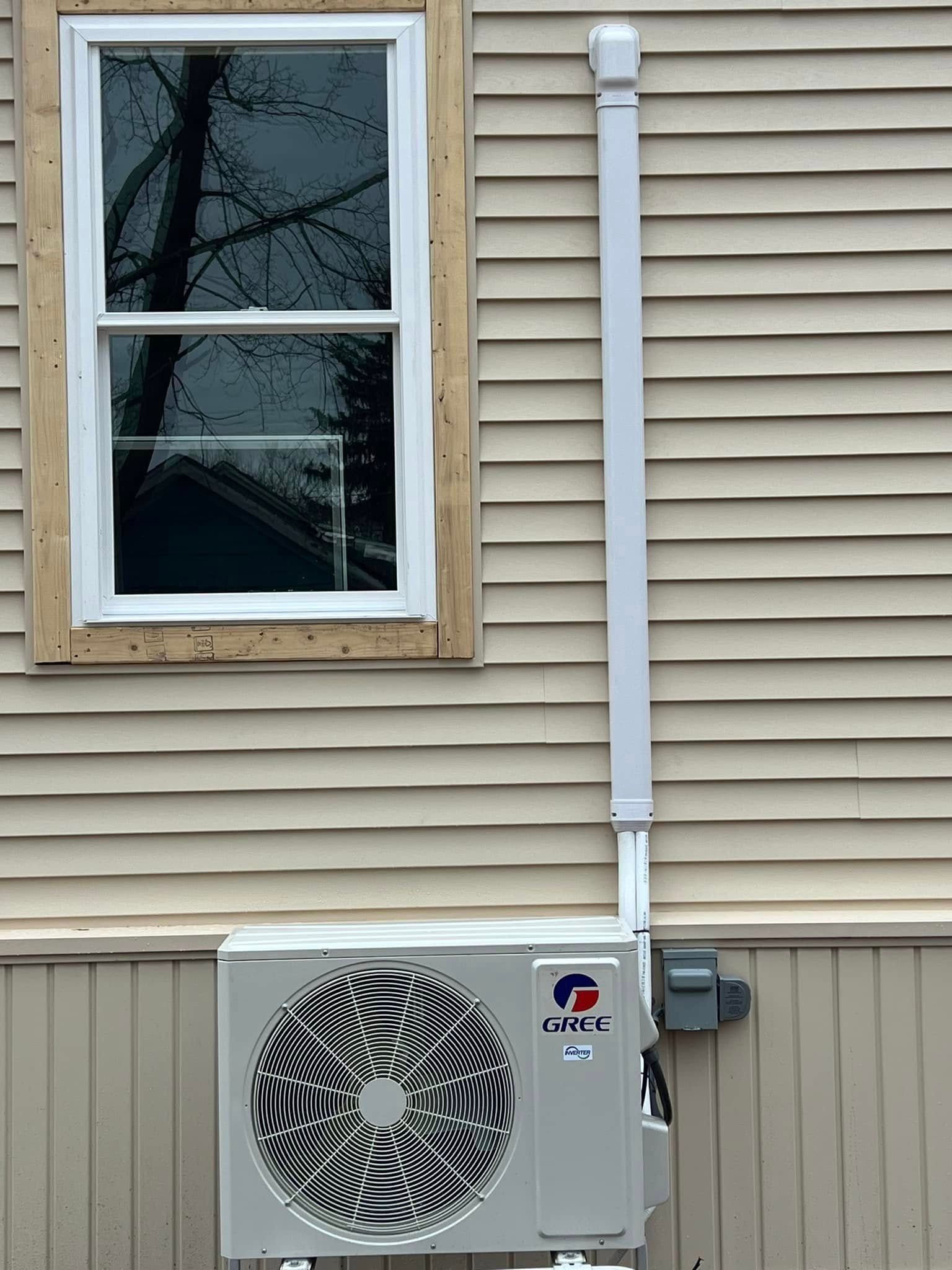 A white air conditioner is mounted on the side of a house next to a window.