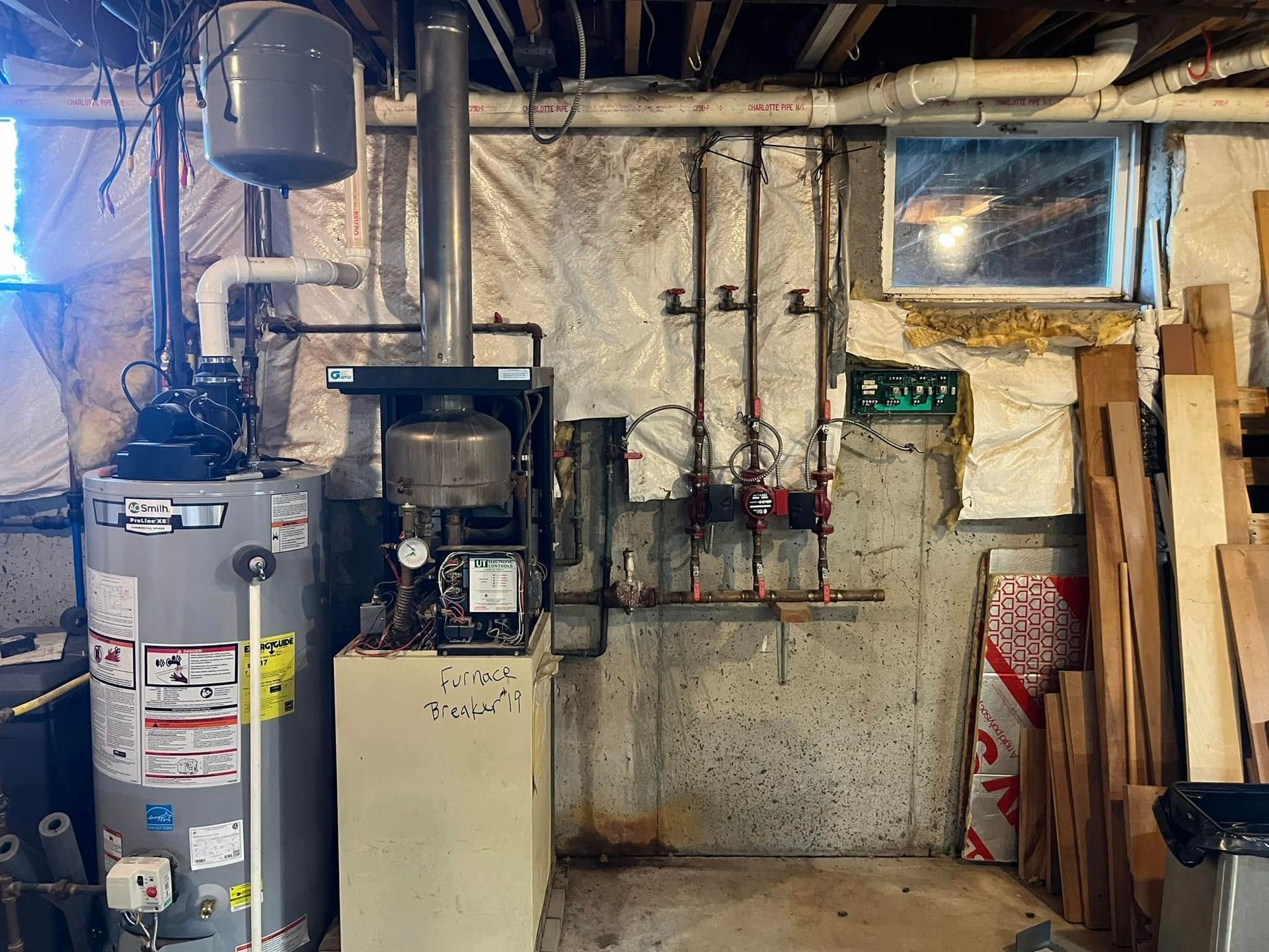 A boiler is sitting in a basement next to a window.