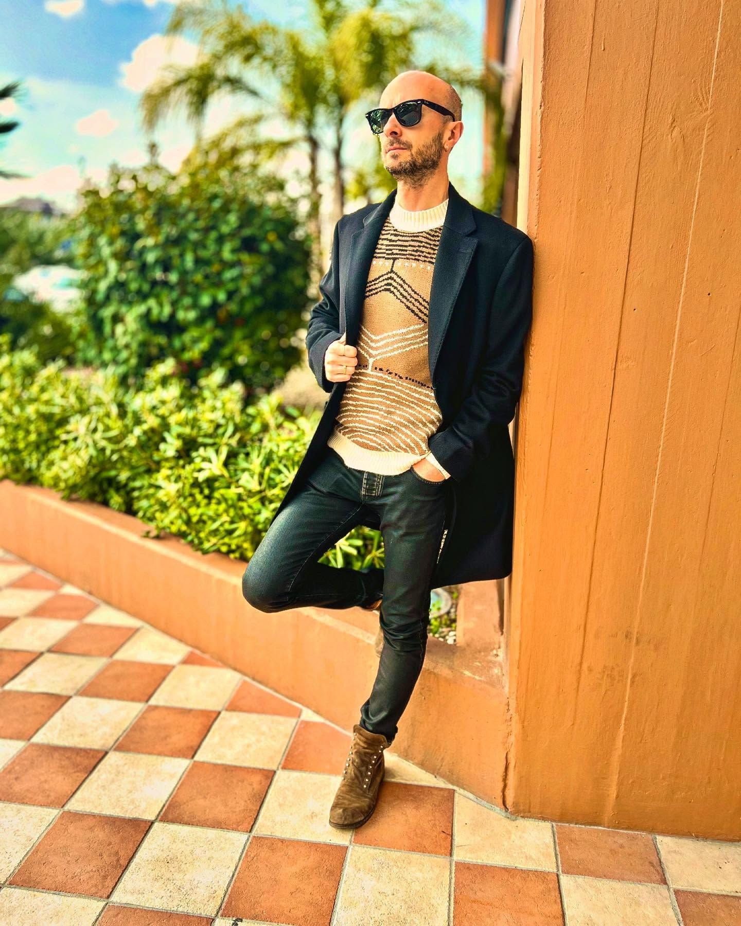 Un uomo appoggiato a un muro che indossa occhiali da sole e un cappotto.