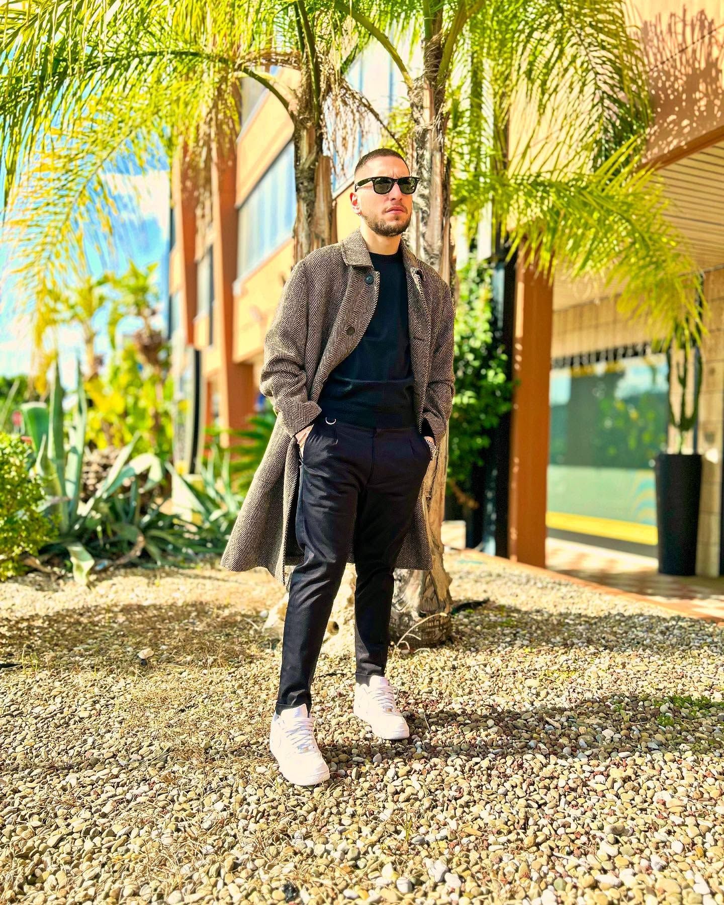 Un uomo che indossa un cappotto e occhiali da sole è in piedi davanti a un edificio.