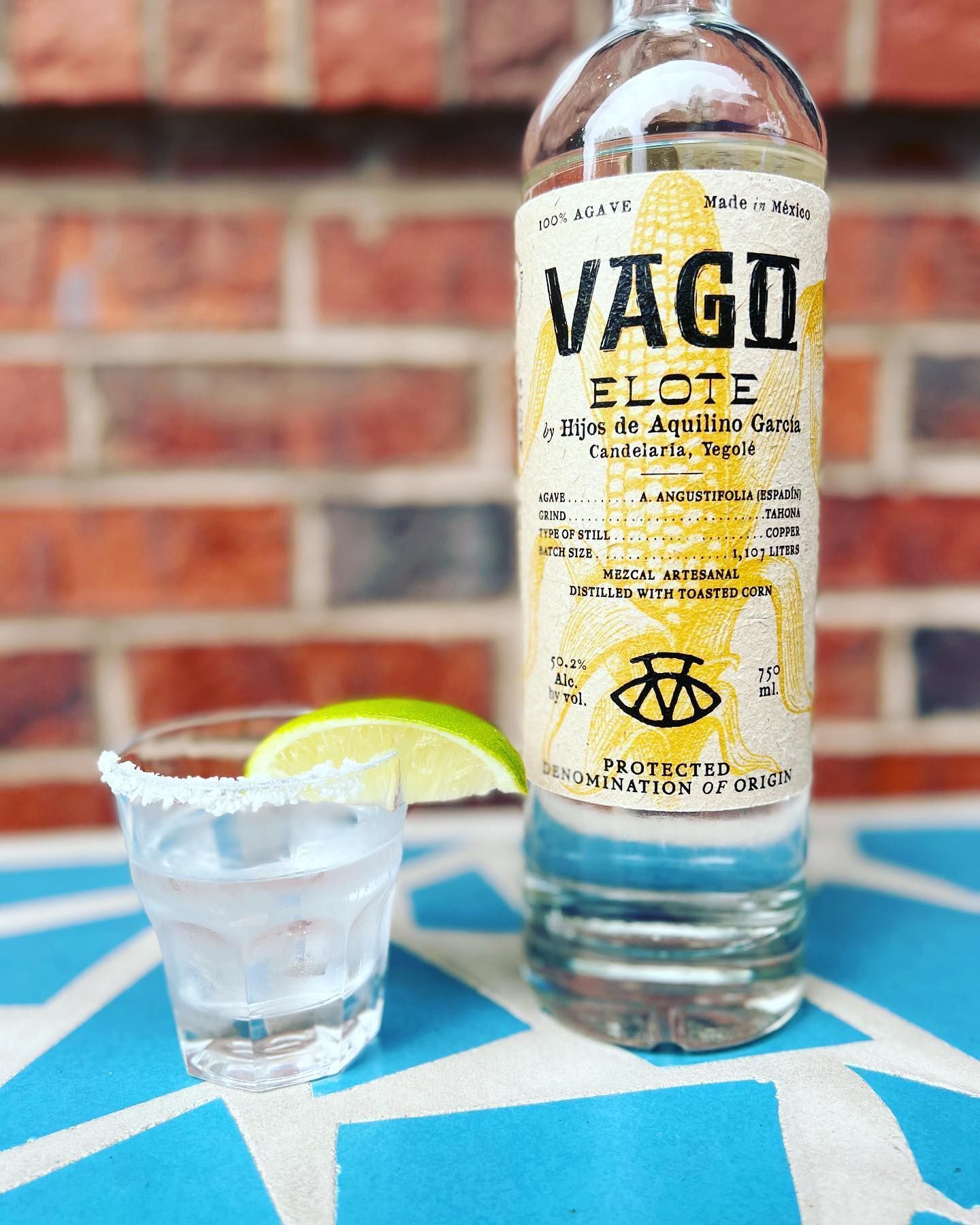 A bottle of vago elote tequila next to a shot glass on a table.