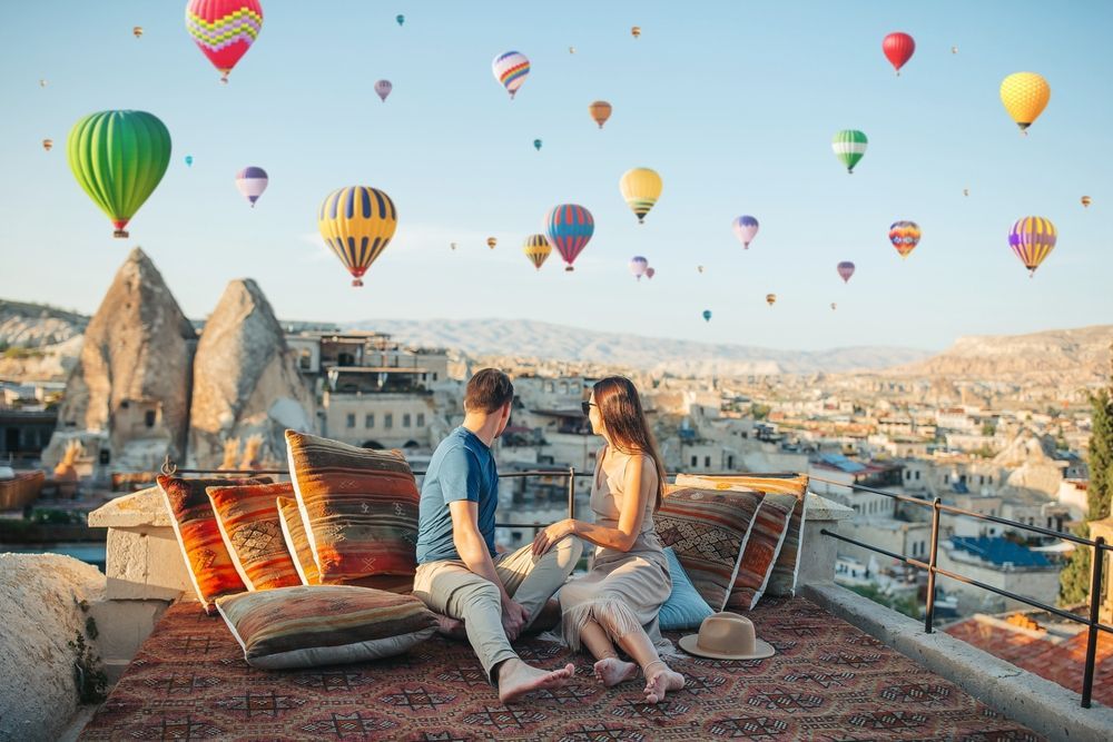 A man and a woman are sitting on the roof of a building with hot air balloons flying in the background.
