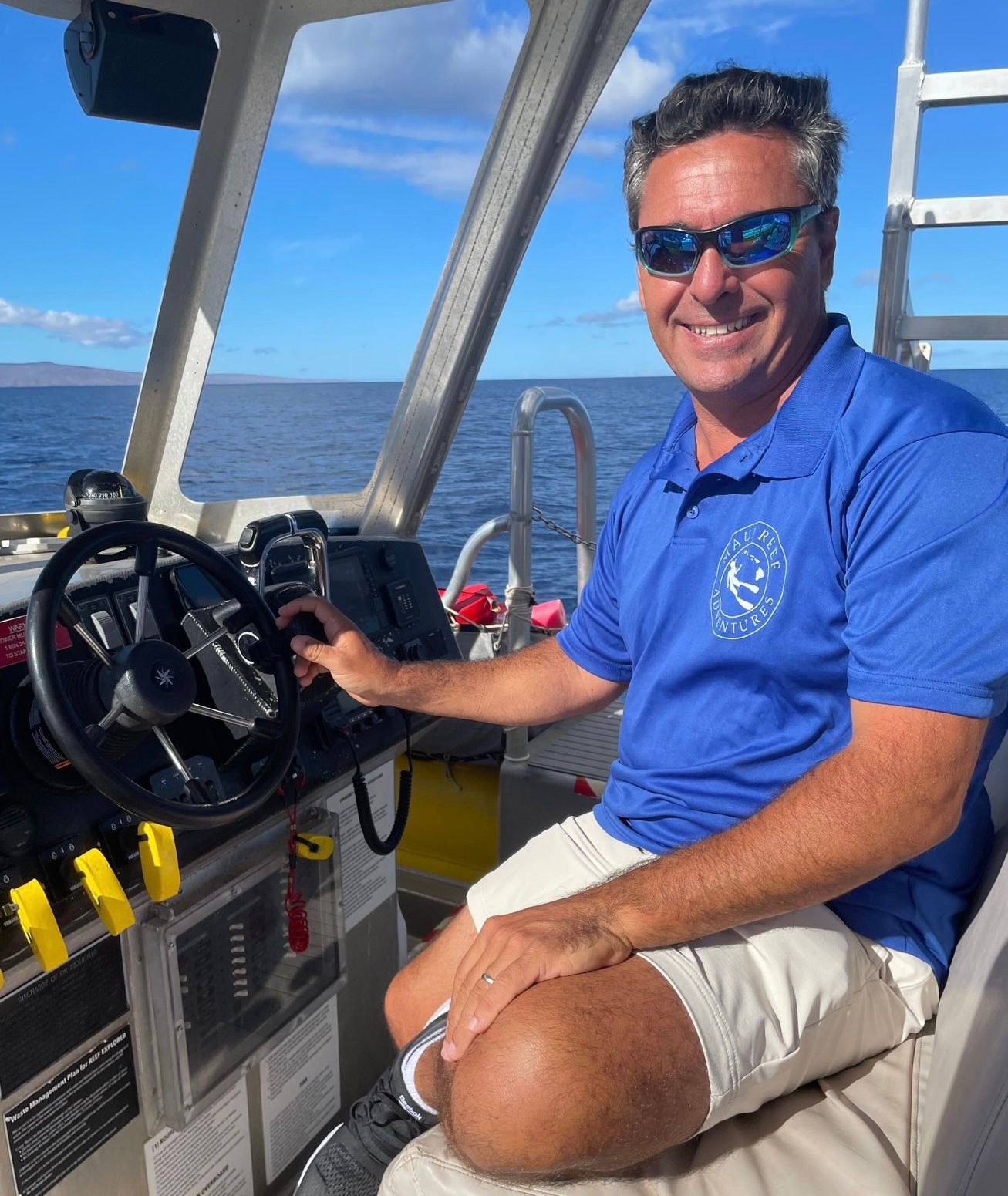 Man steering a boat, smiling, wearing sunglasses and blue polo shirt, ocean background.