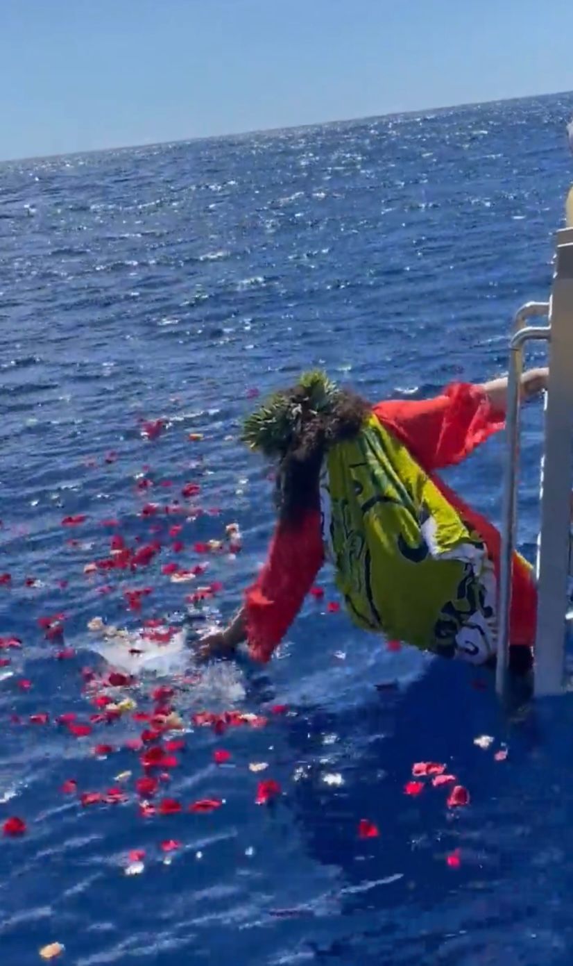 Person in red robes leans over boat, scattering red flower petals into the ocean.