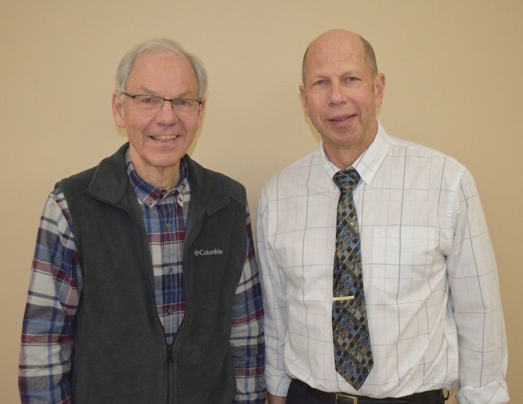 Two men standing side by side indoors, smiling at the camera, one in a plaid shirt and vest, the other in a white shirt and tie