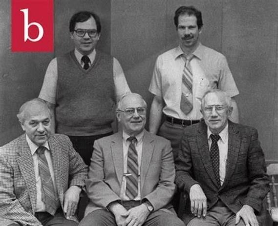 Five men posing in a black-and-white group portrait, with a red square logo in the upper left corner