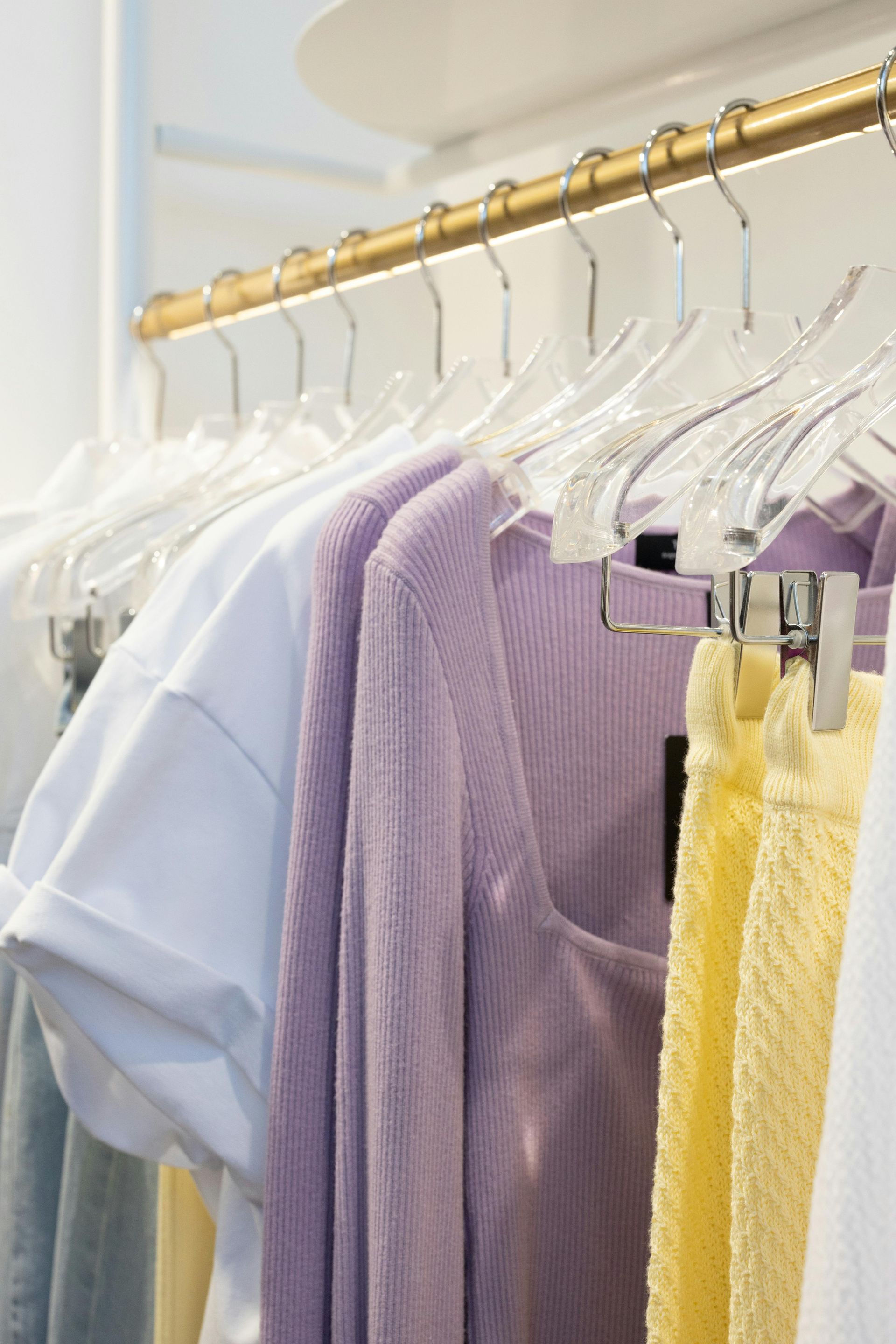 Clothes on clear hangers hanging from a gold rack, featuring a white top, a lilac sweater, and a textured yellow skirt.