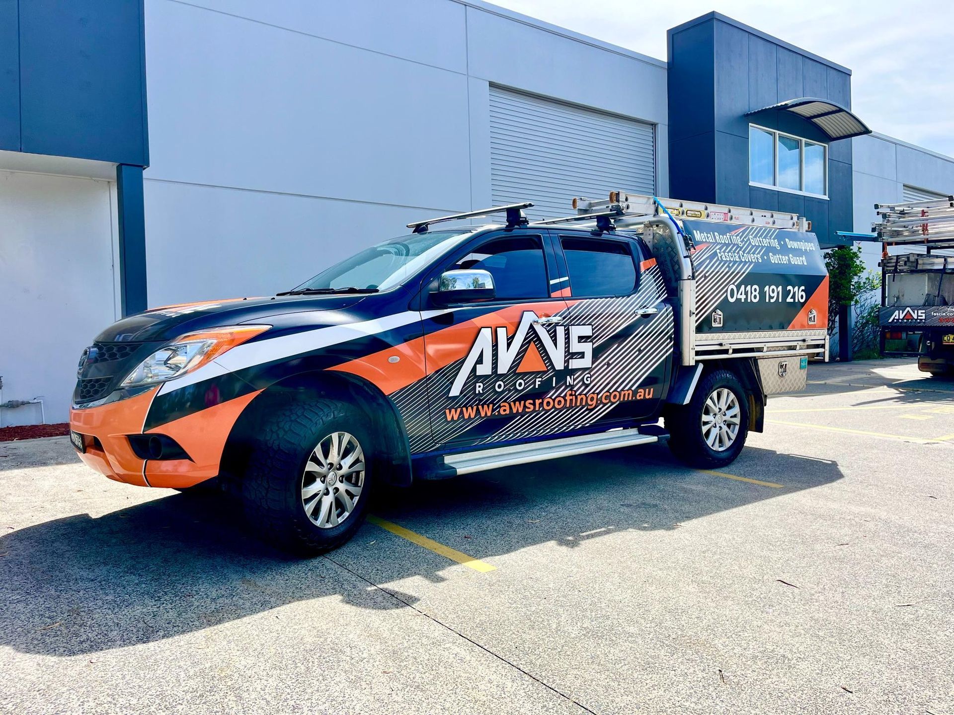 A pickup truck with orange and black graphics, parked outside a building, with a ladder on top.