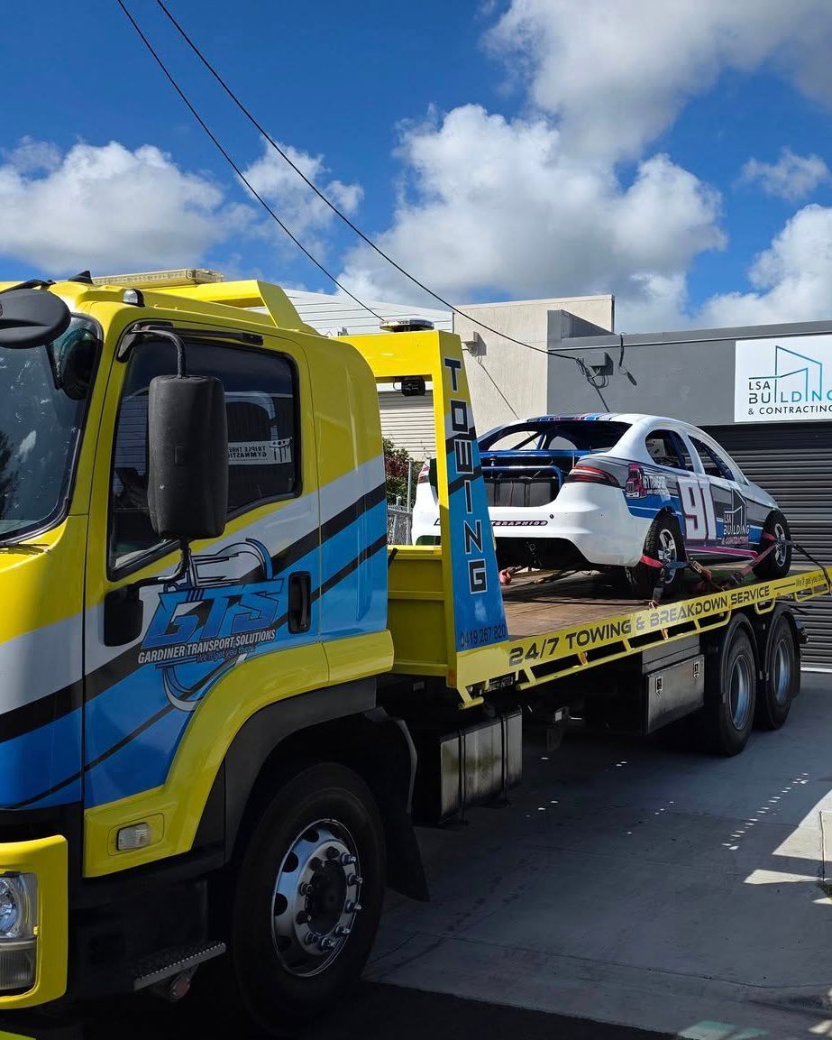 Yellow and blue tow truck hauling a white race car under a blue sky — GTS - Gardiner Transport Solutions in Berserker, QLD