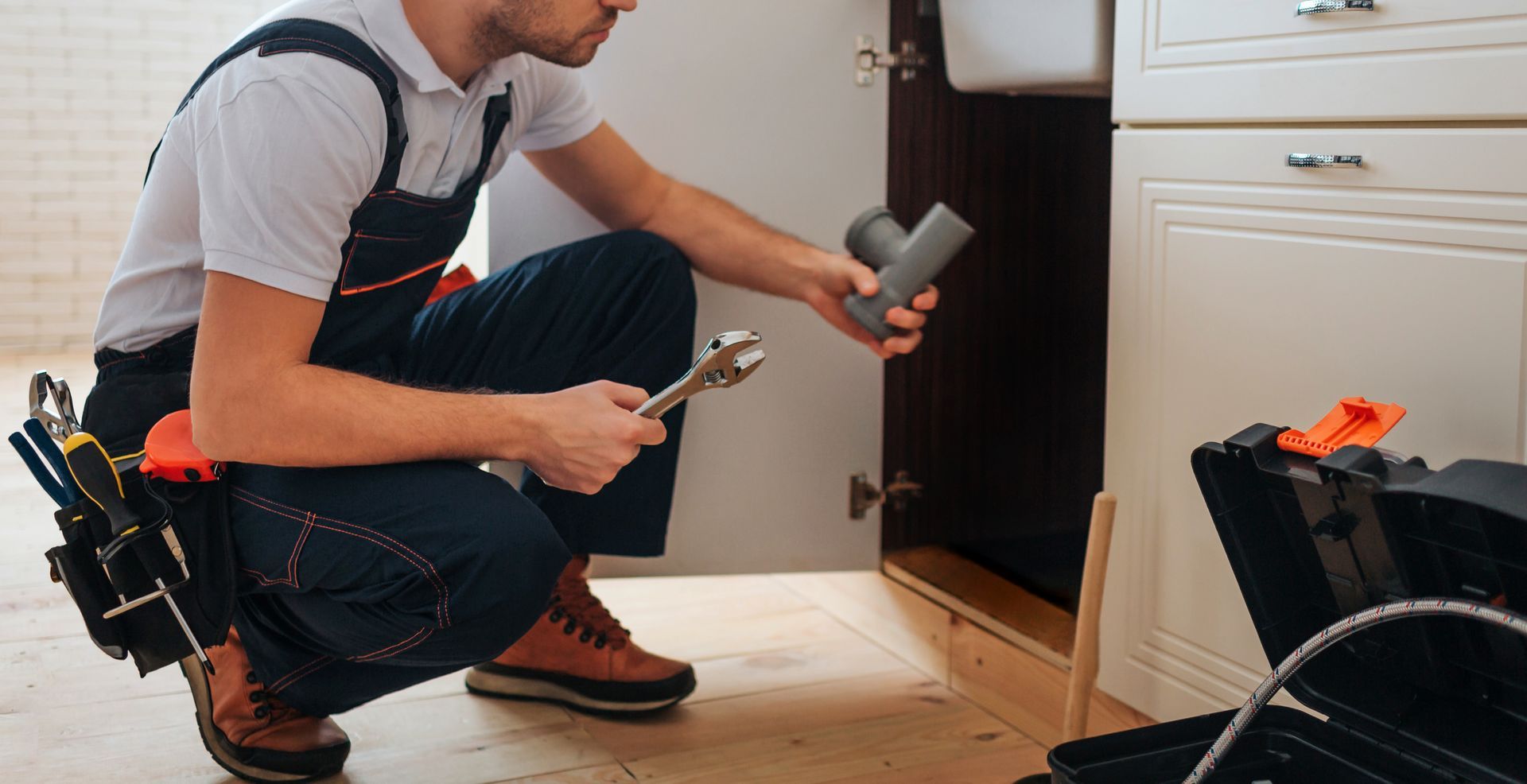 Plumber crouches under sink holding pipe and wrench beside open cabinet.