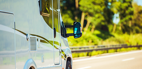 White RV traveling on a road with green trees in the background. Side mirror reflects the surroundings.