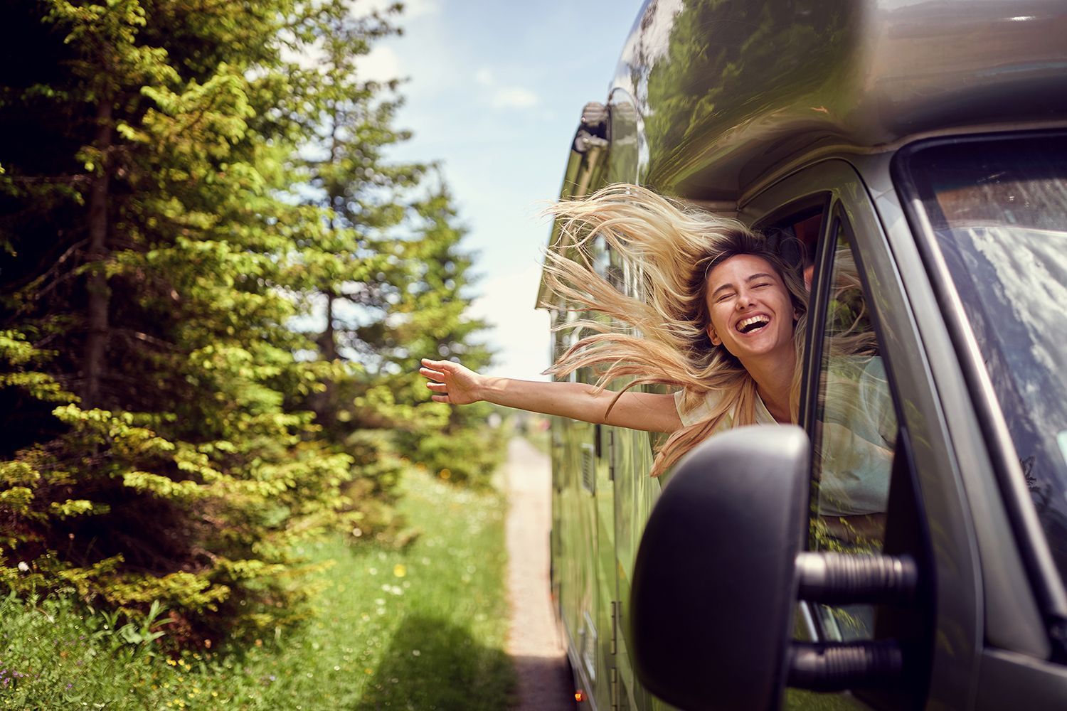 A person leans out of a moving van with hair blowing in the wind along a forest road.