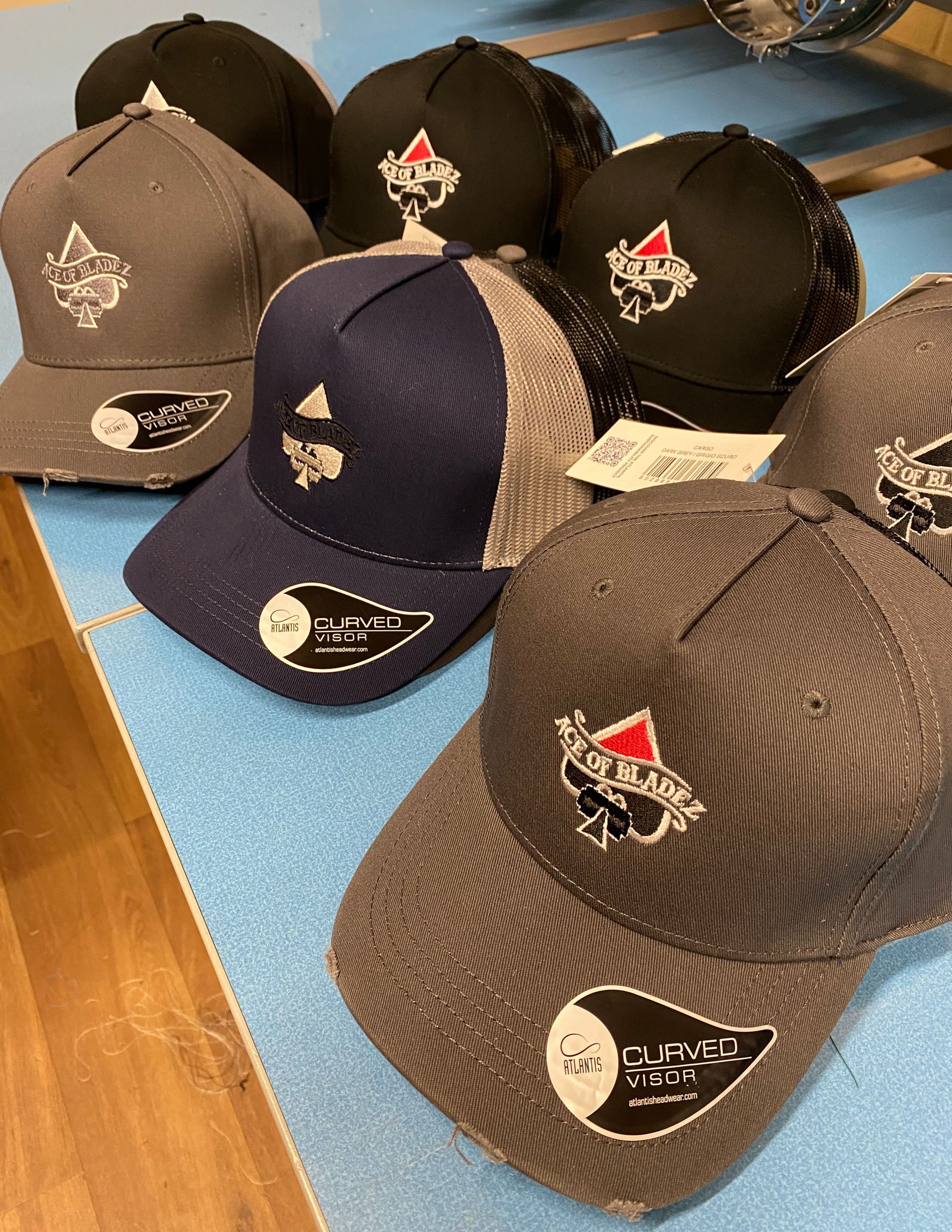 Several baseball caps with embroidered Ace of Bladez logo, in various colors, displayed on a blue surface.