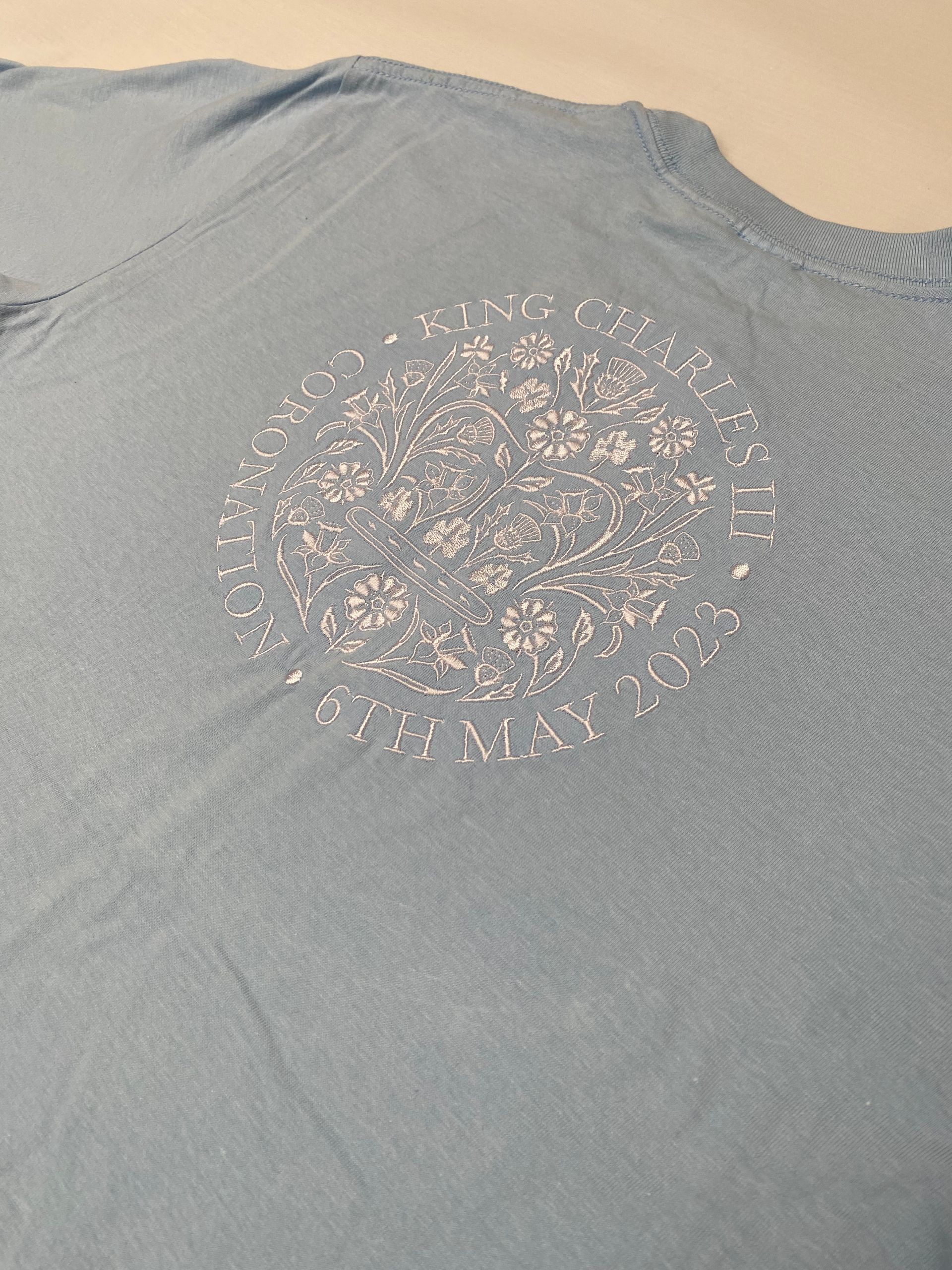 Light blue t-shirt with a embroidered white floral design, including text King Charles III Coronation 6th May 2023