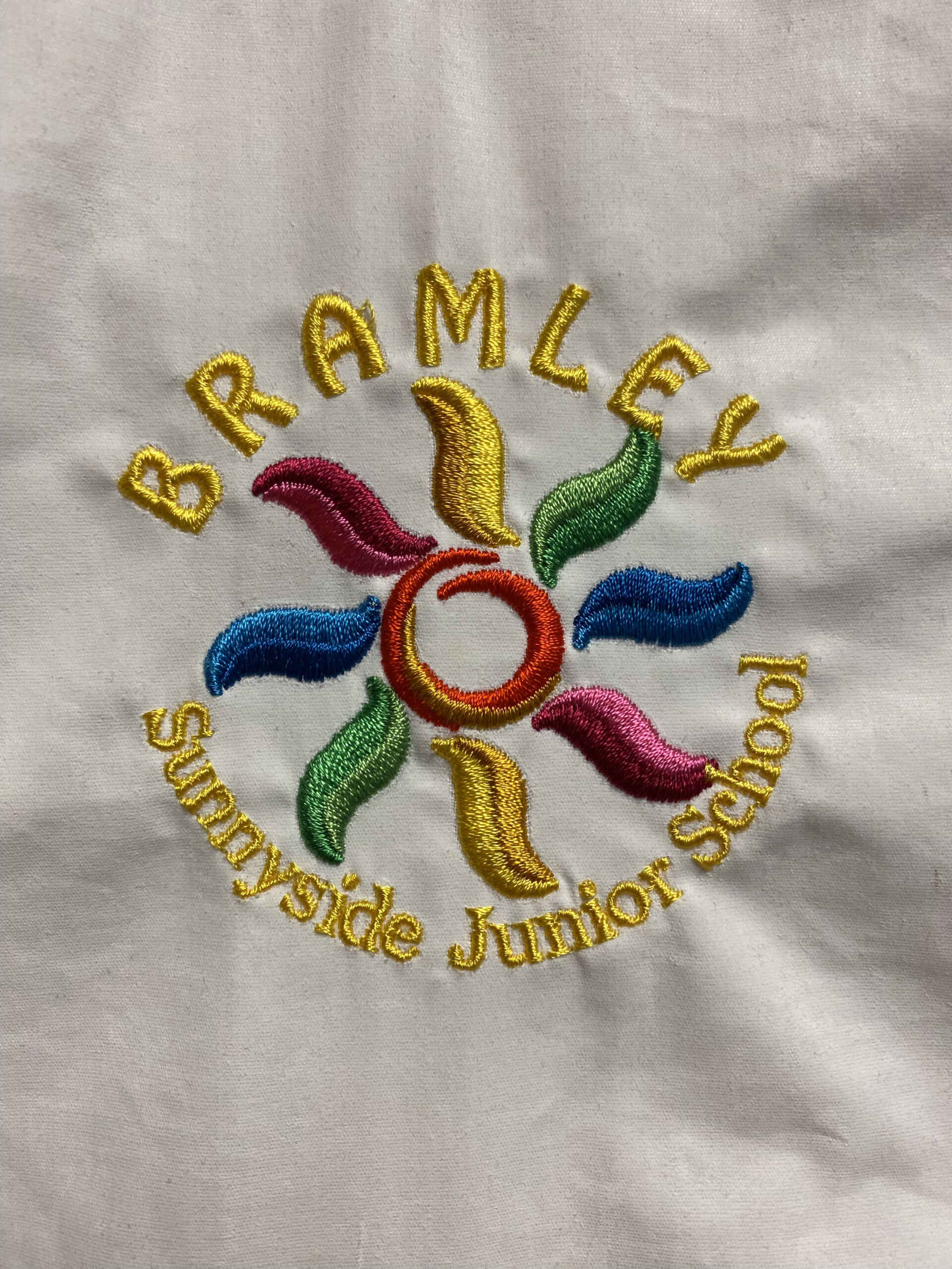 Embroidered logo of Bramley Sunnyside Junior School. Features a colourful sun and text in gold thread on white fabric.