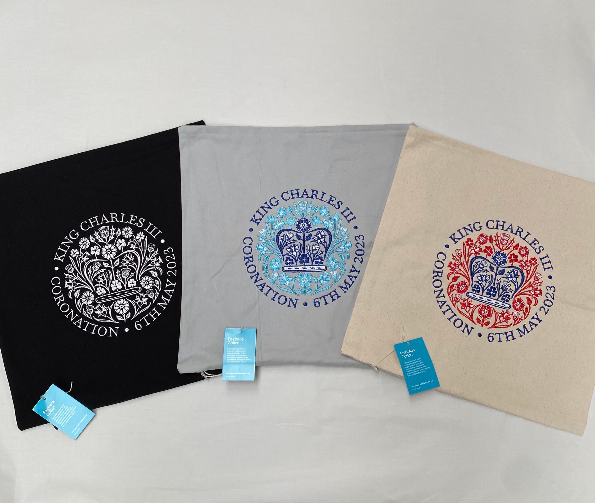 Three pillow cases in black, grey, and beige, each with a embroidered coronation emblem in different colours.