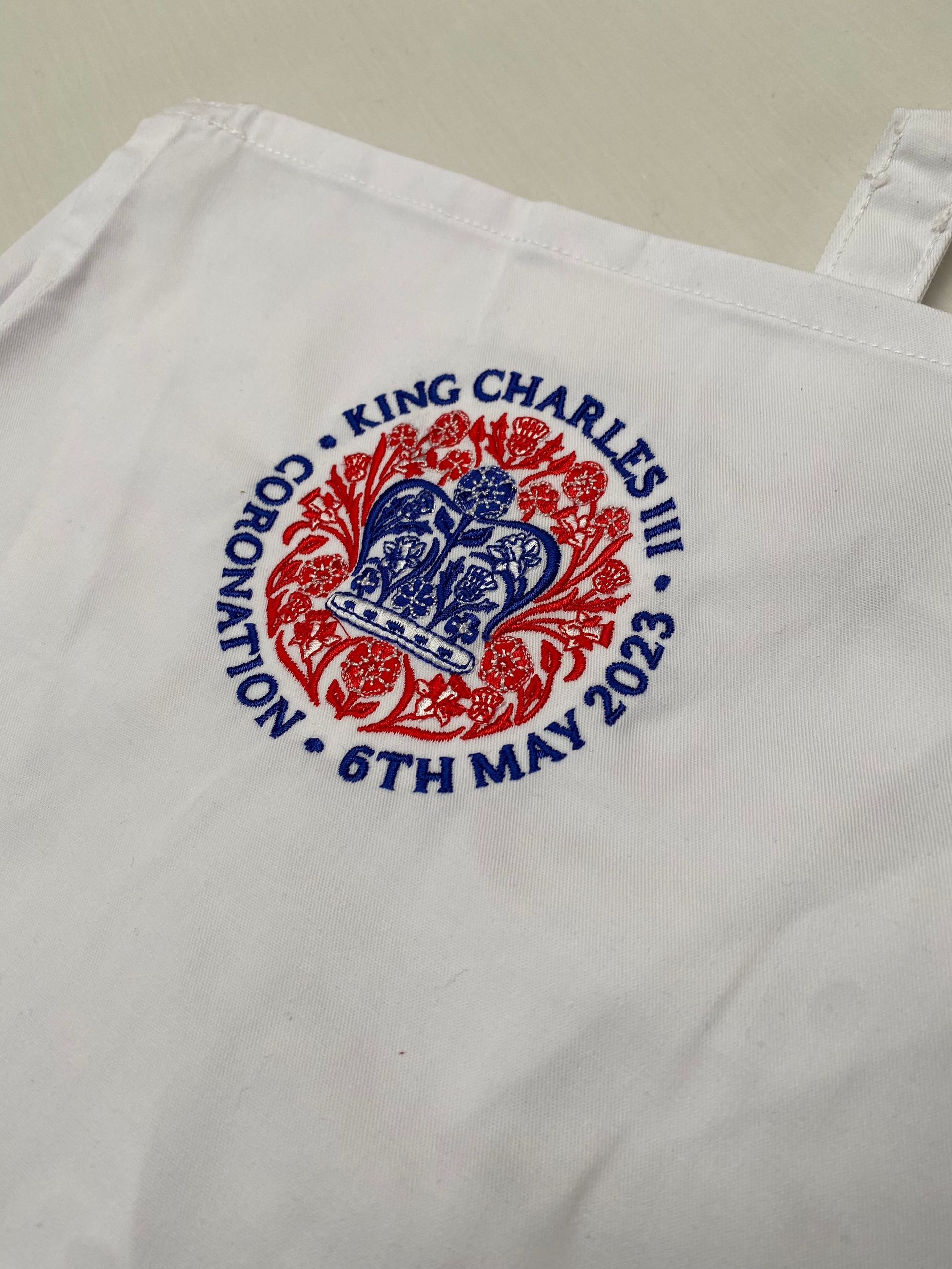 White apron with a King Charles III coronation embroidered emblem in red and blue, dated May 6, 2023.