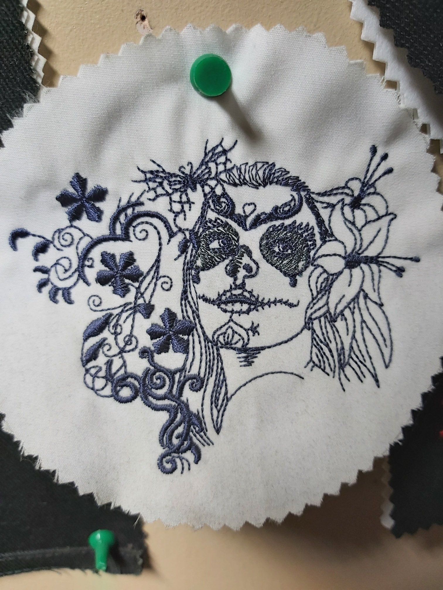 A circular white fabric swatch embroidered with a dark blue sugar skull design, pinned to a surface.