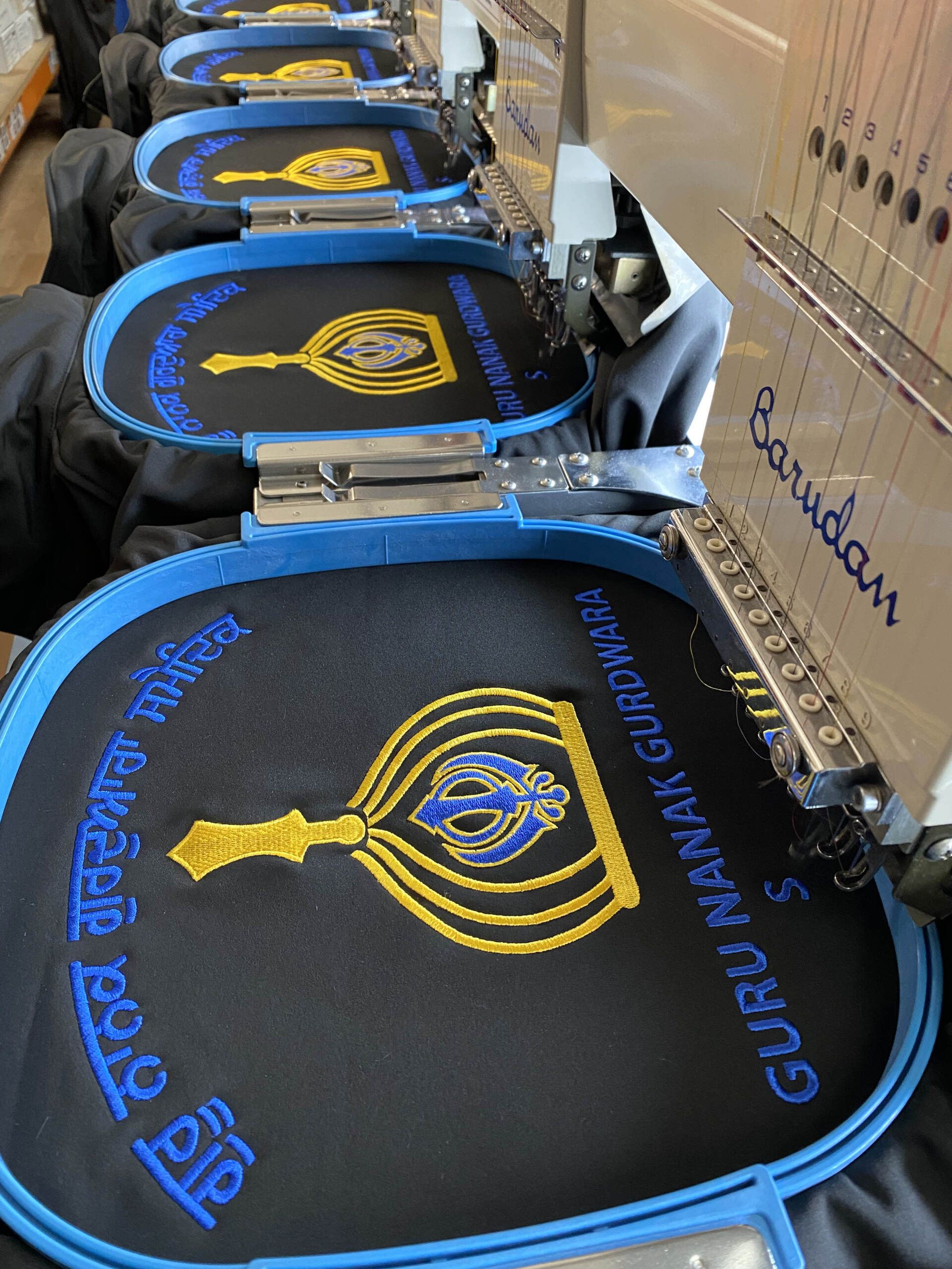 Embroidery machine in operation, stitching the GNG design onto black fabric, labeled