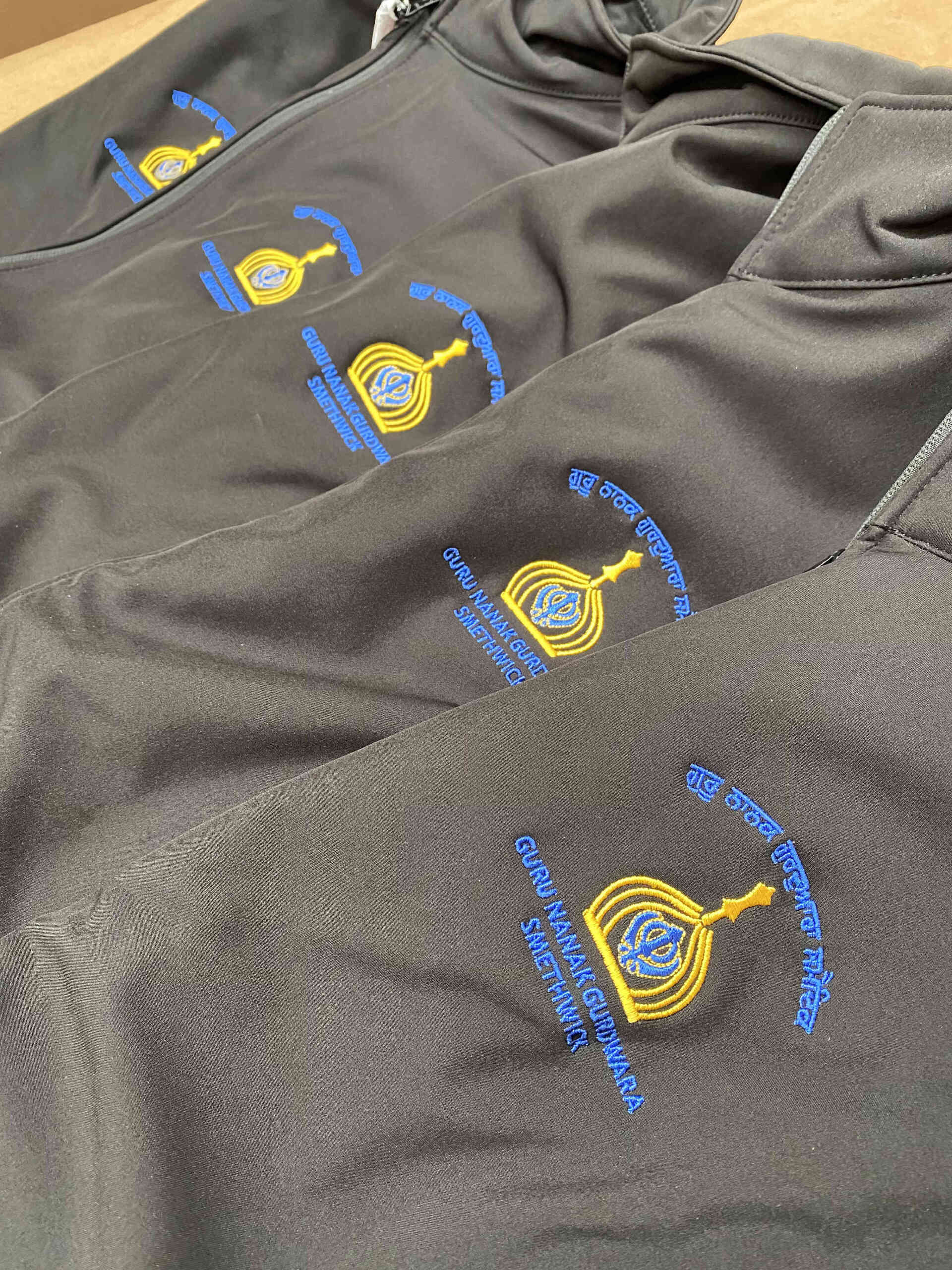 Four black jackets with embroidered Guru Nanak Gudwara logos: a gold with blue accents and text embroidered to left chest
