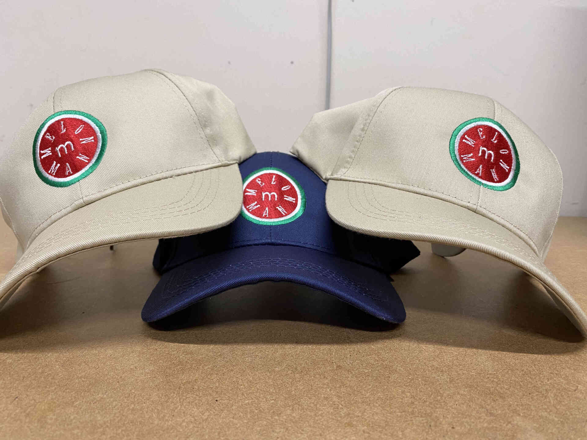 Three baseball caps with a the melon man embroidered logo, two tan and one blue, sit on a surface.